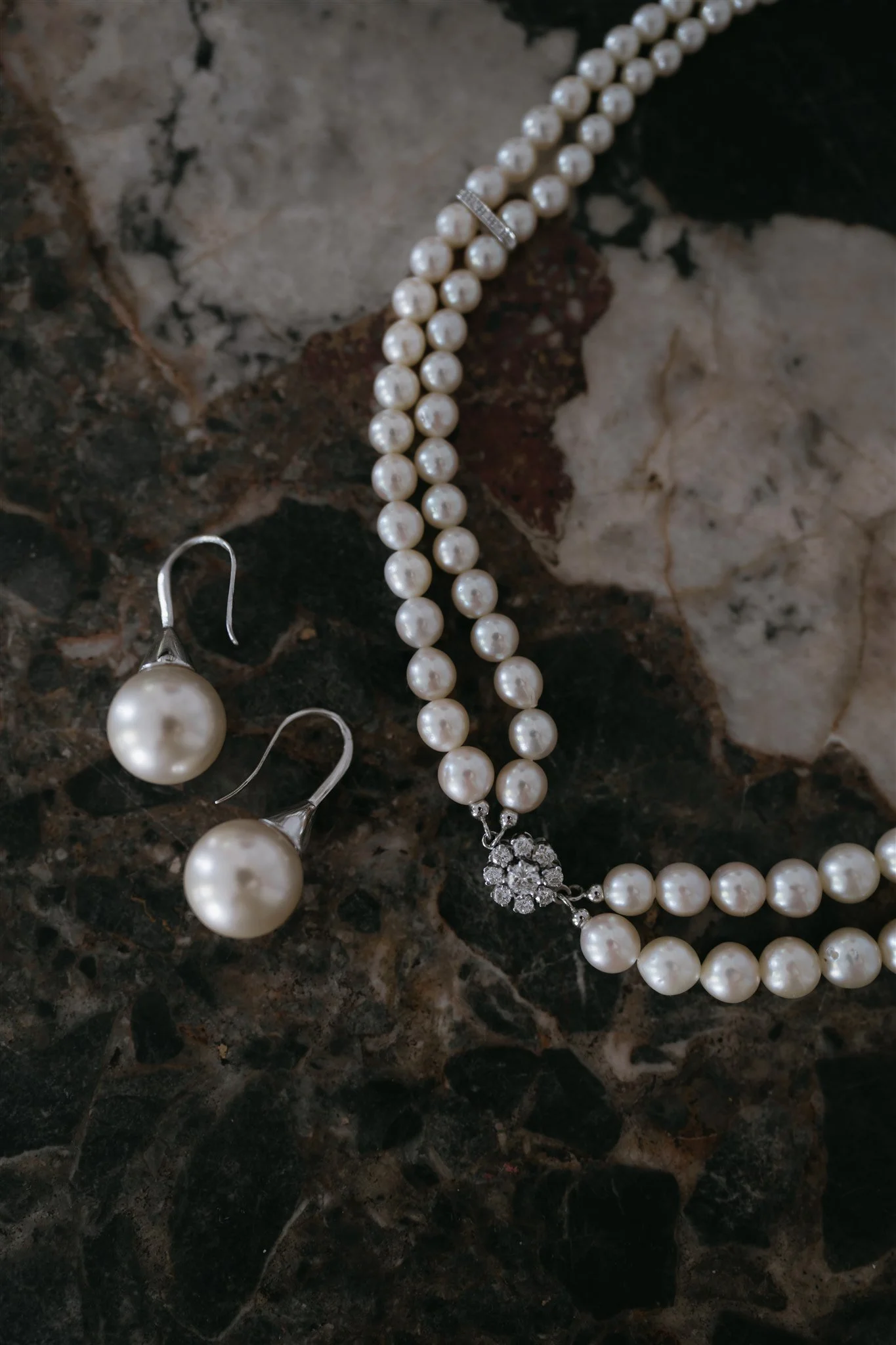 A close-up of elegant bridal jewelry featuring a pearl necklace with a delicate floral clasp and matching pearl drop earrings, displayed on a luxurious marble surface.