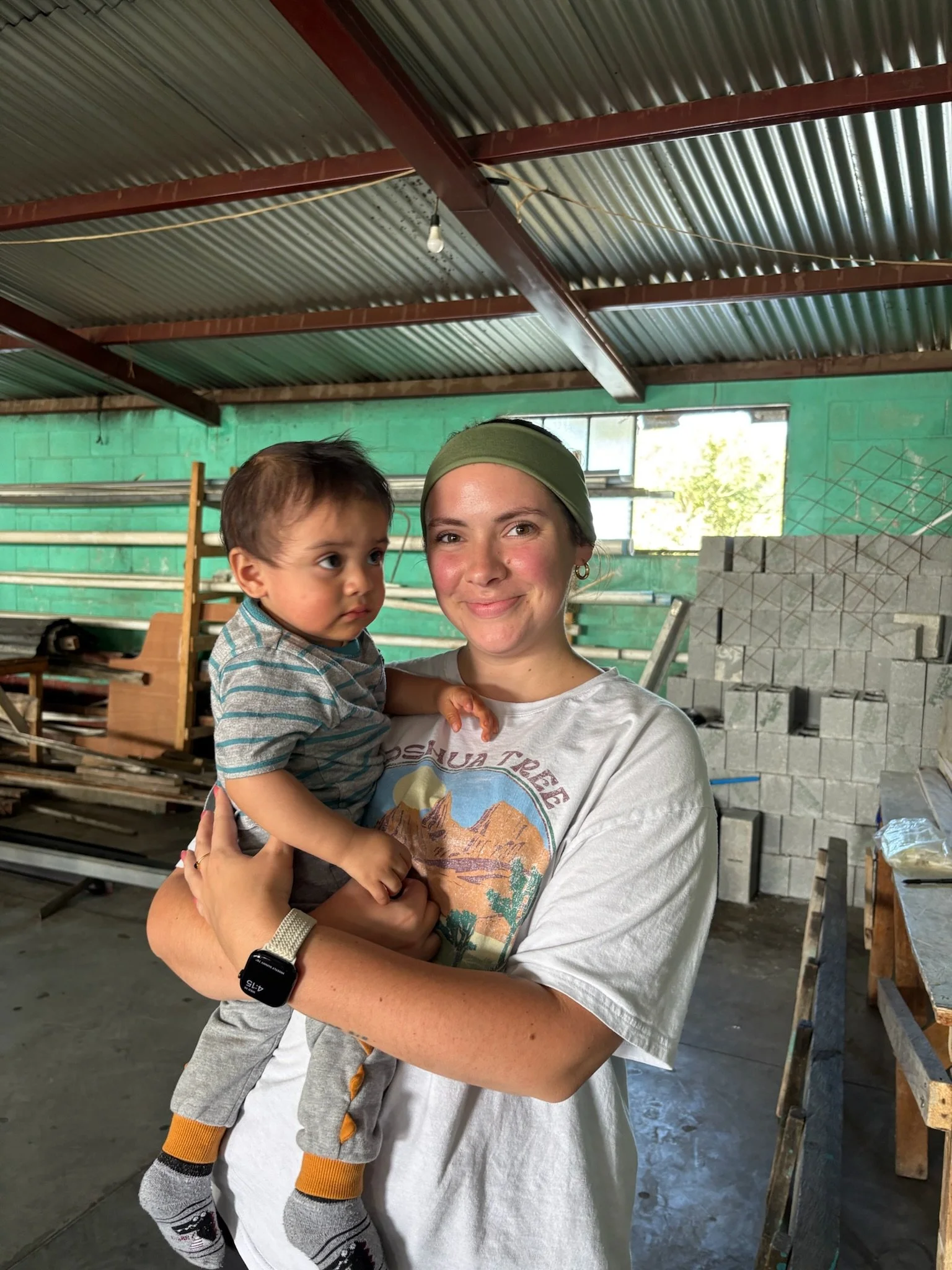 A woman with light skin, wearing a green headband, a white T-shirt with a desert mountain graphic, and a smartwatch, is holding a young boy with light skin, brown hair, wearing a striped T-shirt, gray pants, and colorful socks, inside a workshop with