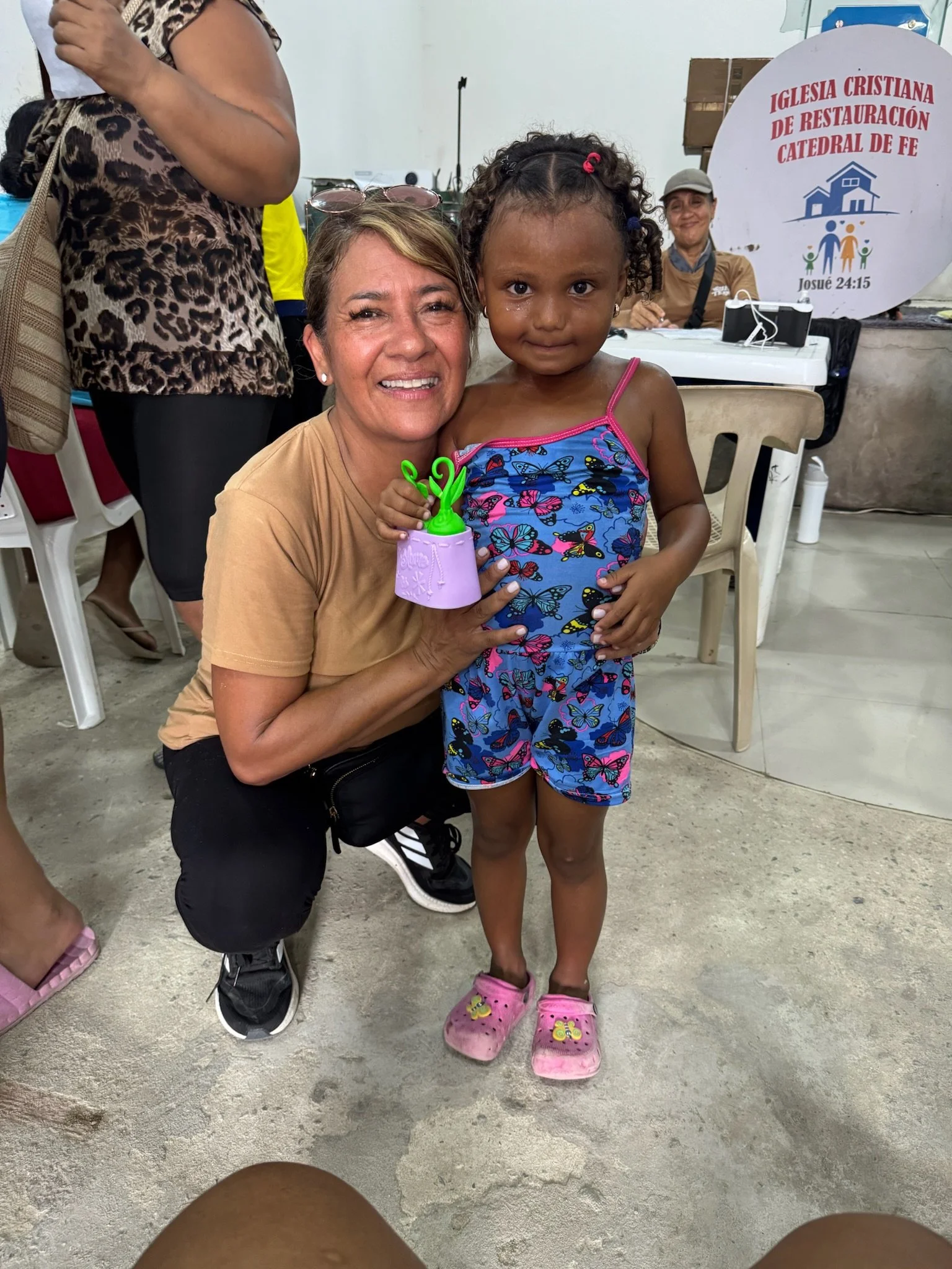 A woman crouching and smiling next to a young girl with butterfly pajamas. The woman is holding a toy, and they are inside a room with chairs and a sign in the background that reads 'Iglesia Cristiana de Restauración Catedral de Fe.'