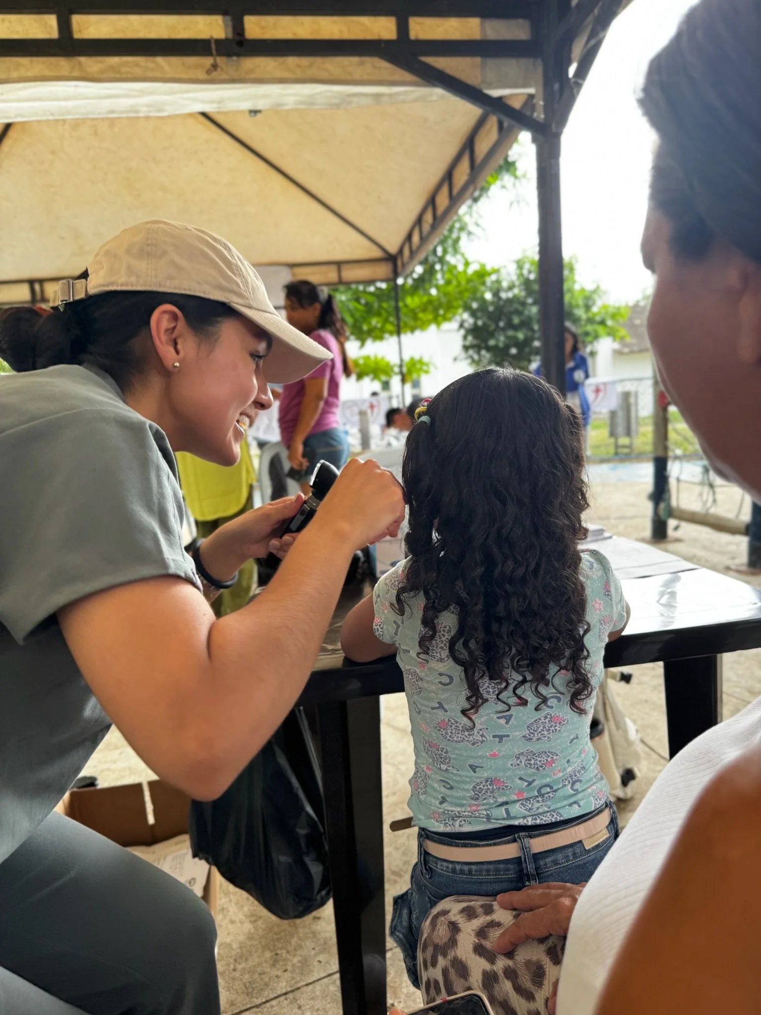 A woman with a beige hat is taking close-up photos of a young girl with curly hair at an outdoor event under a canopy, with other people and trees in the background.