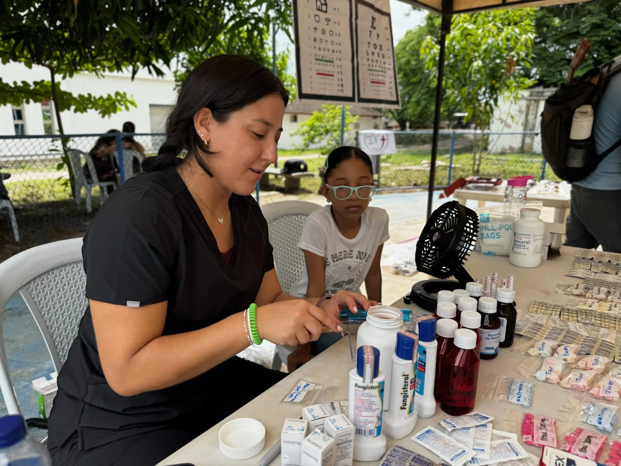 A woman and a girl at an outdoor table with medical supplies, including bottles and packets of medication, under a canopy with trees in the background.