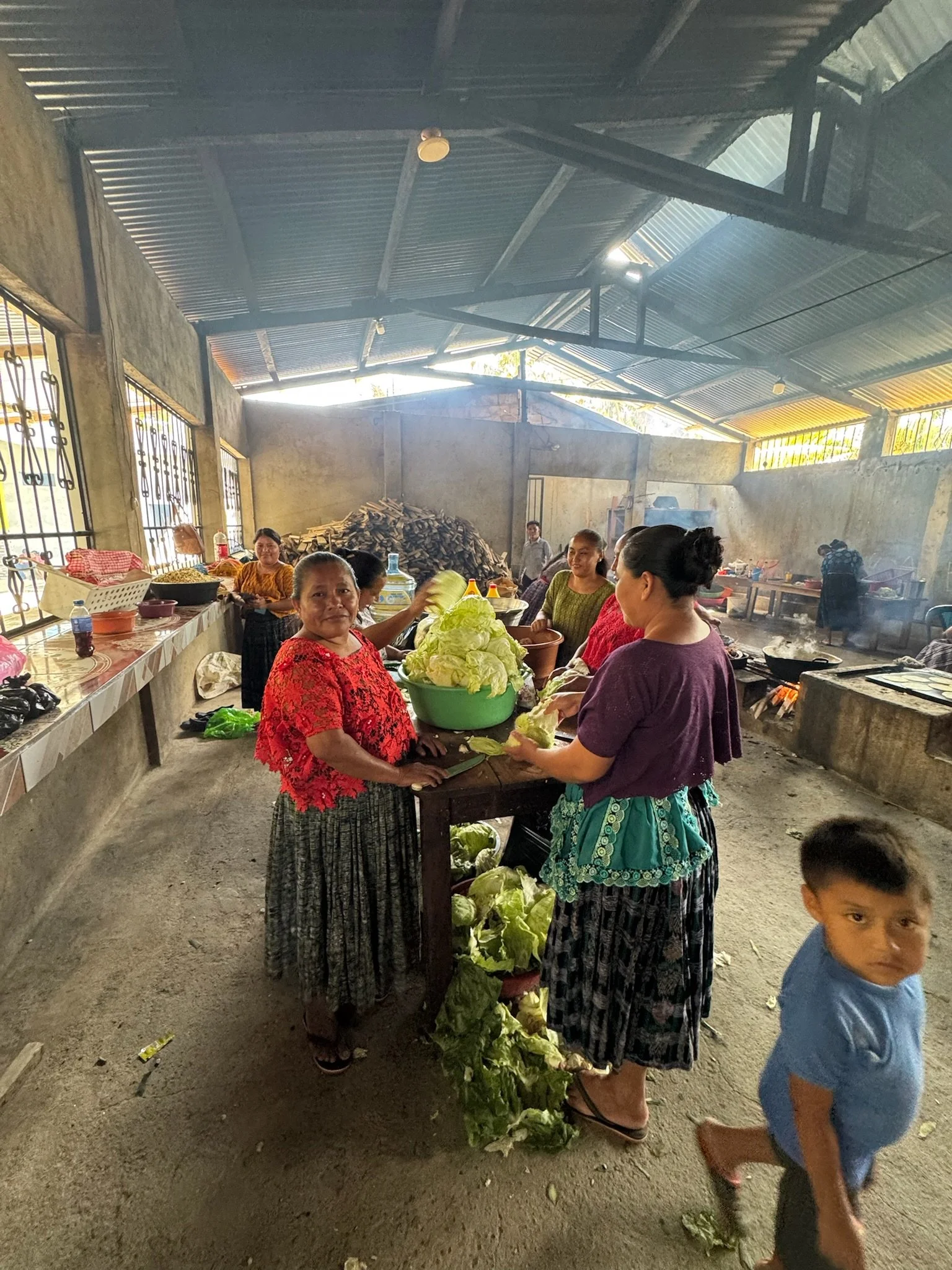 Women preparing and selling vegetables, including cabbages, inside a rustic market or gathering space, with a young boy in the foreground.