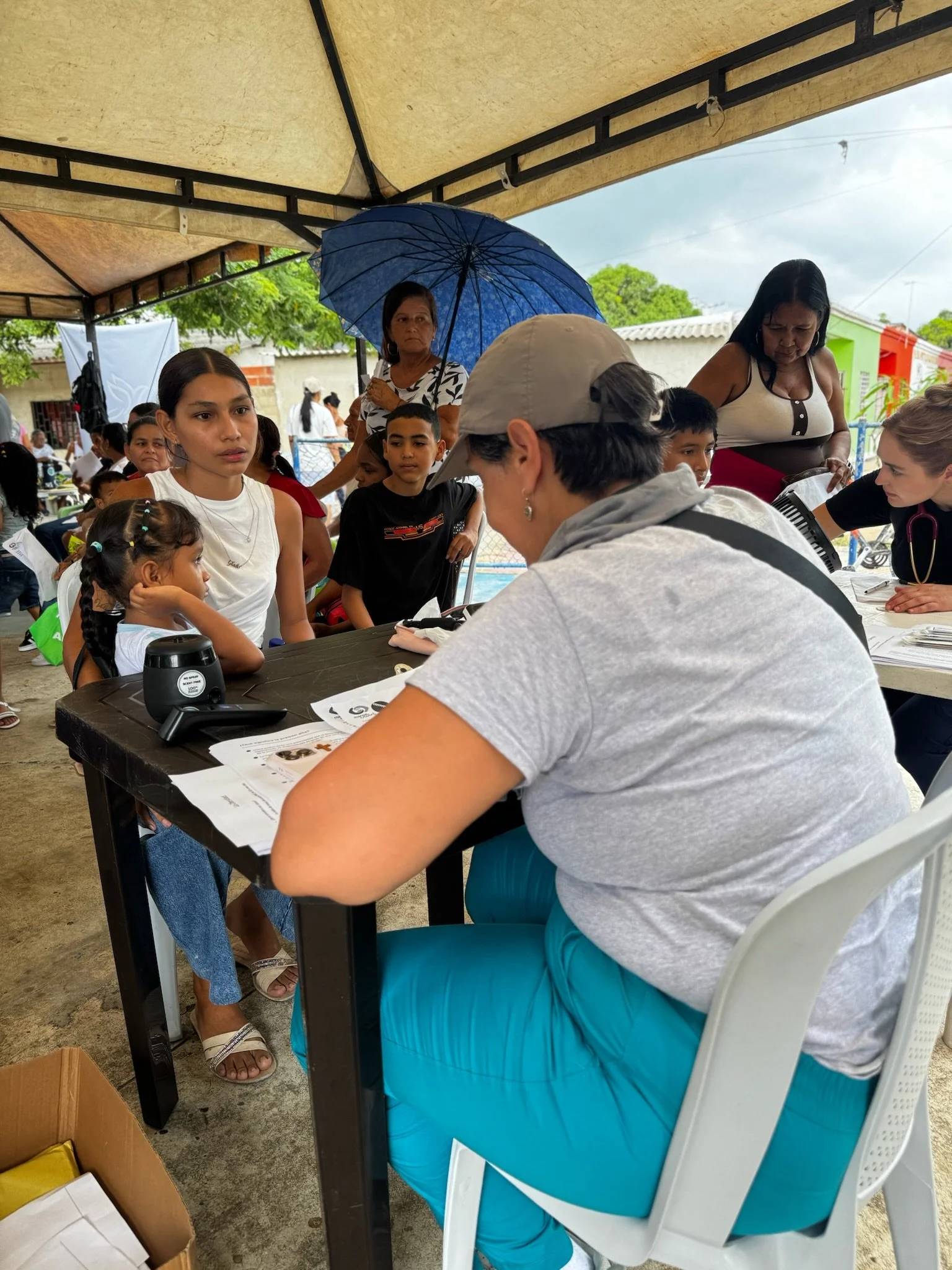 Children and adults at a health clinic or registration booth under a tent, with healthcare workers and a woman holding an umbrella outside.