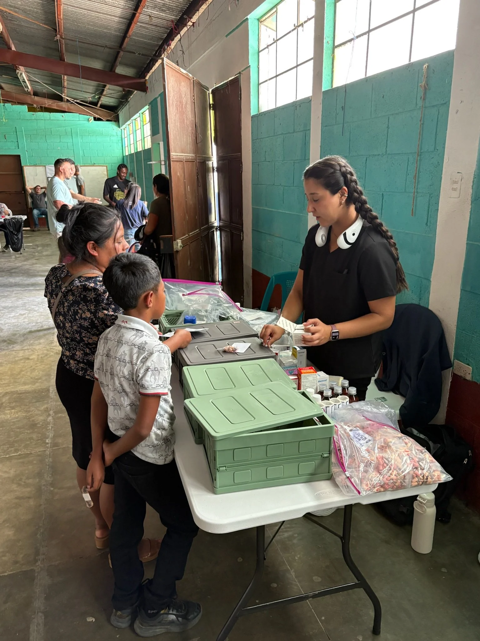 A healthcare worker with braids and black scrubs, wearing headphones, standing behind a table with medical supplies, giving medication to children and adults in a community setting.
