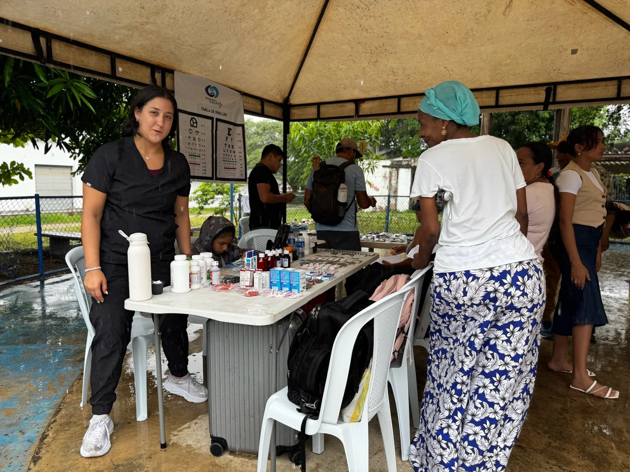 Outdoor health fair with medical supplies and staff, rain-soaked pavement, and several women and children congregating around a table under a canopy.