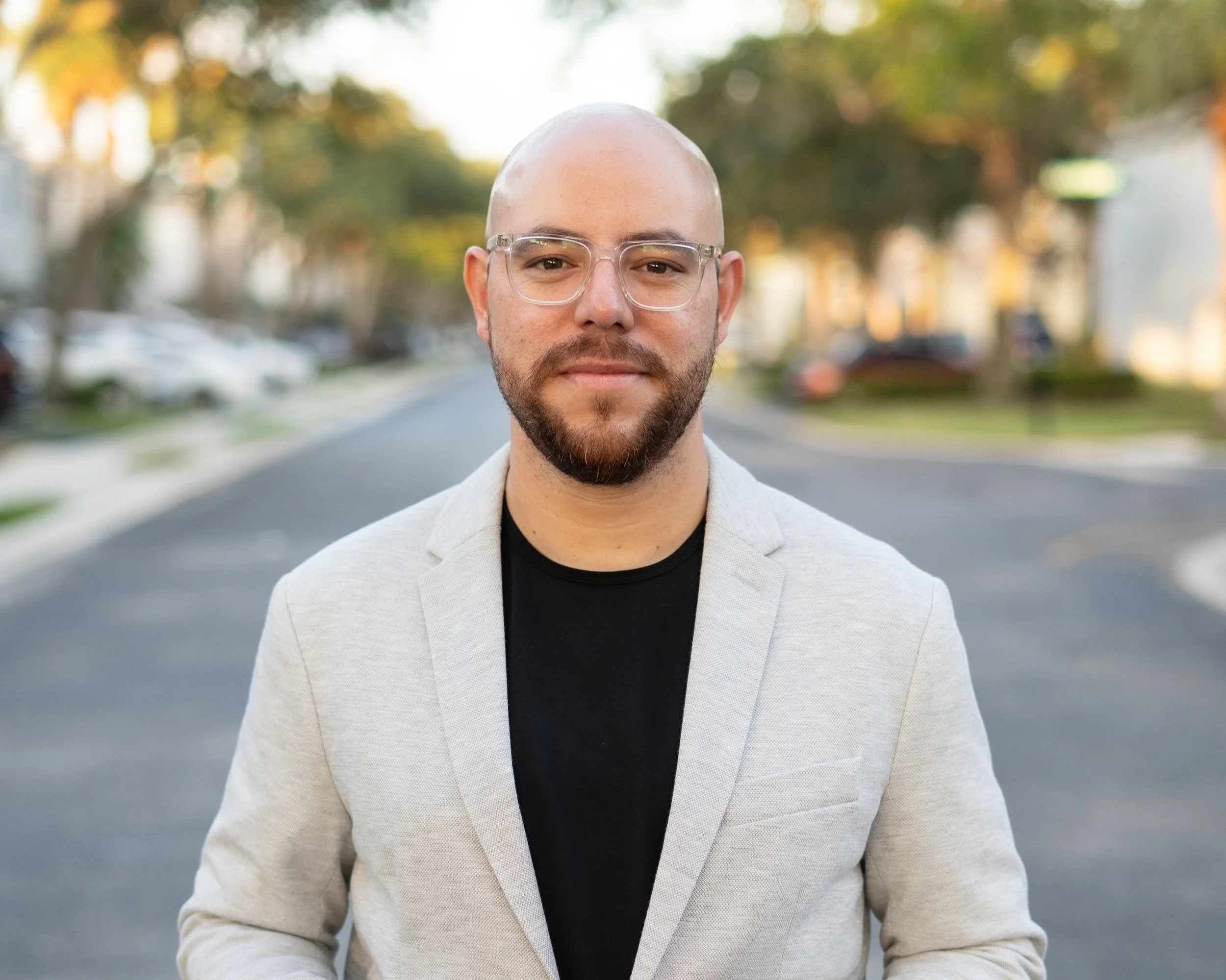 A man with a beard, glasses, and a shaved head standing on a street during daytime with trees and cars in the background.