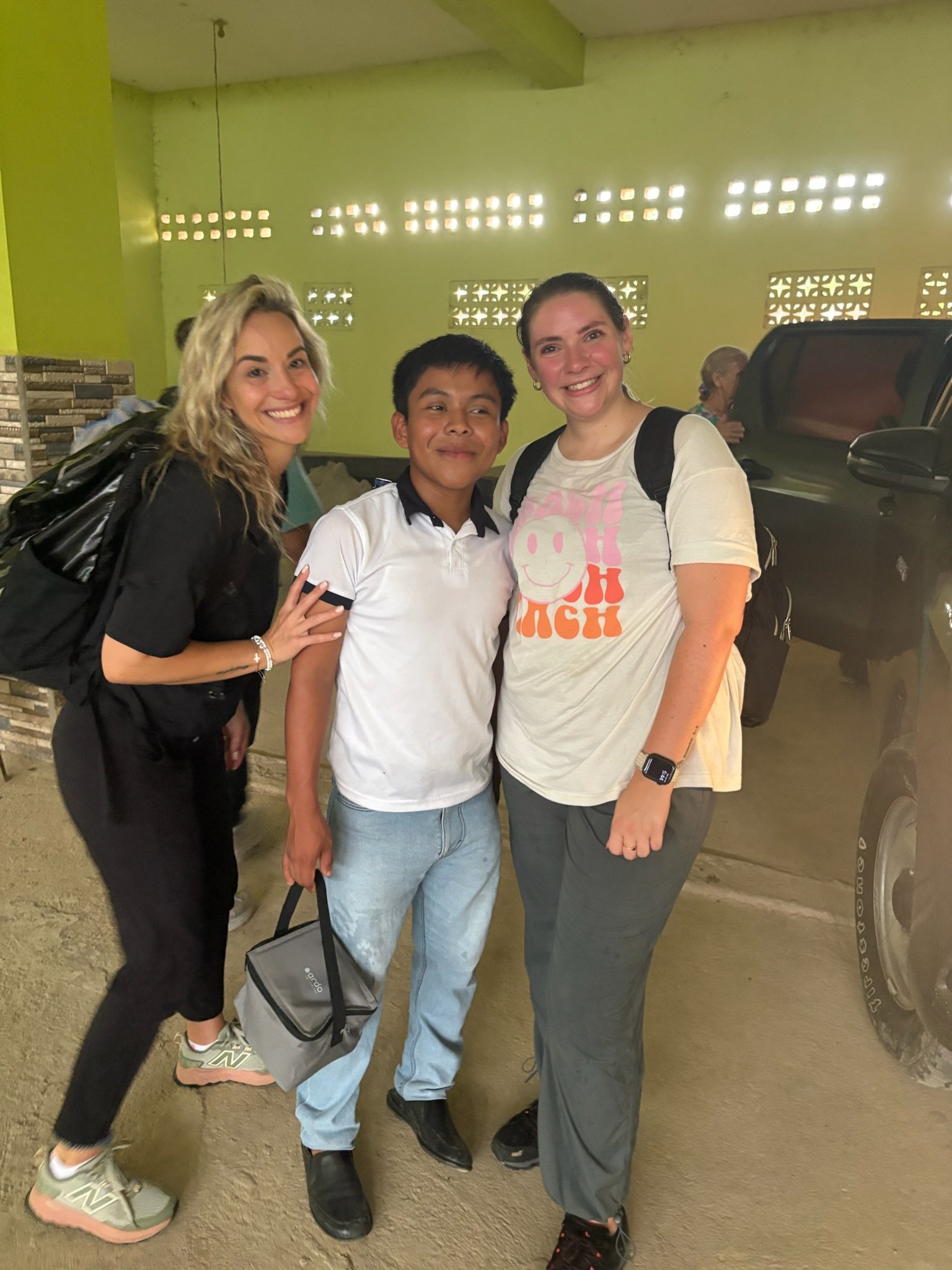 Three people standing together, smiling at the camera, in an indoor space with a green wall and a vehicle in the background.