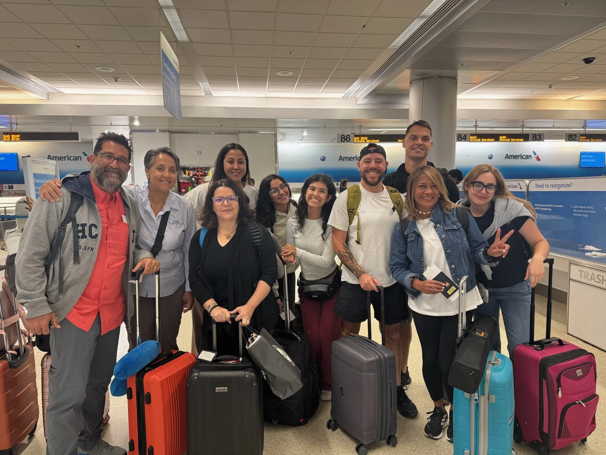 Group of ten diverse travelers standing together at an airport with luggage, smiling for the photo.