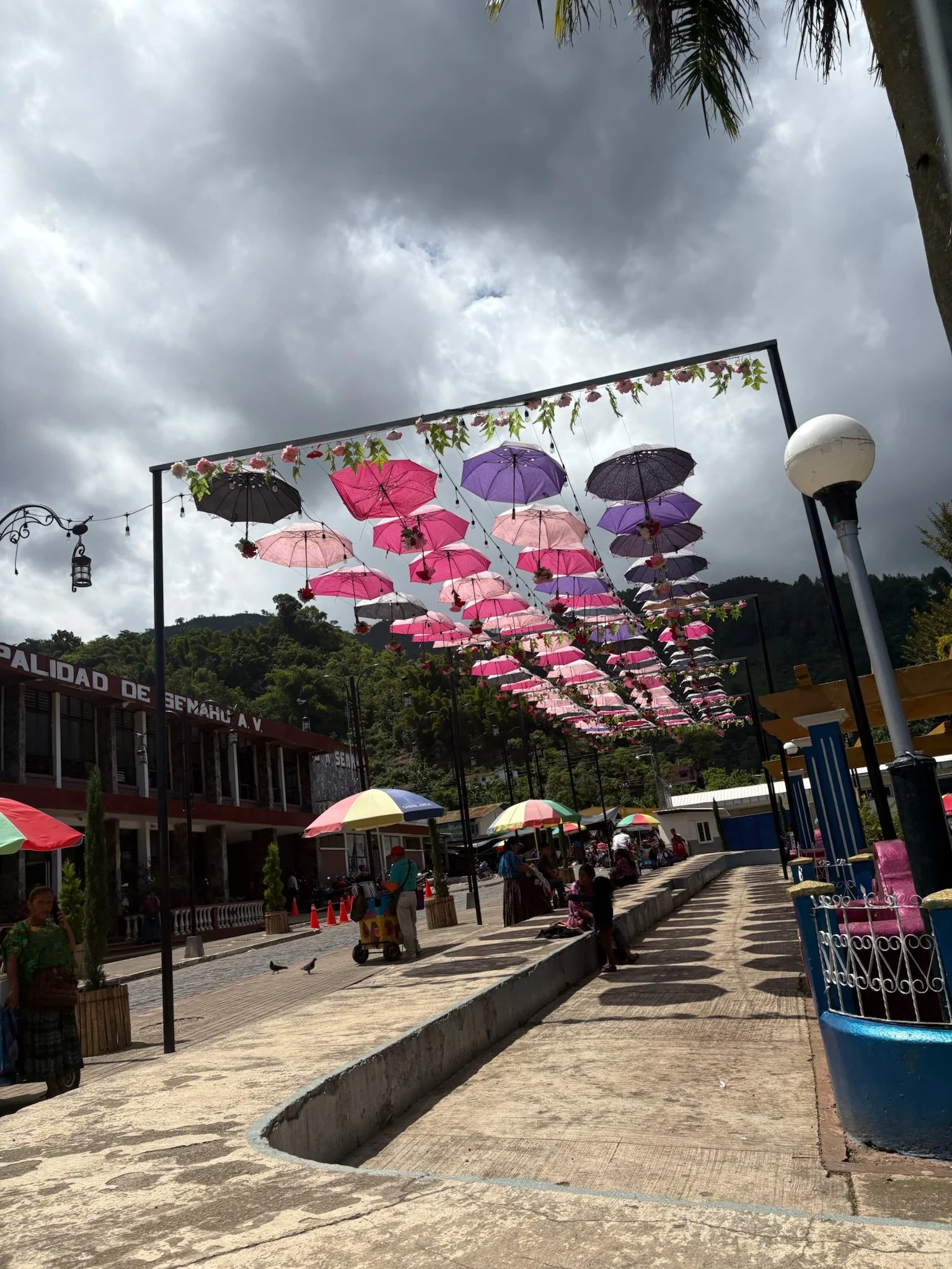 Street scene with hanging umbrellas and people walking, colorful umbrellas, cloudy sky, and mountainous background.