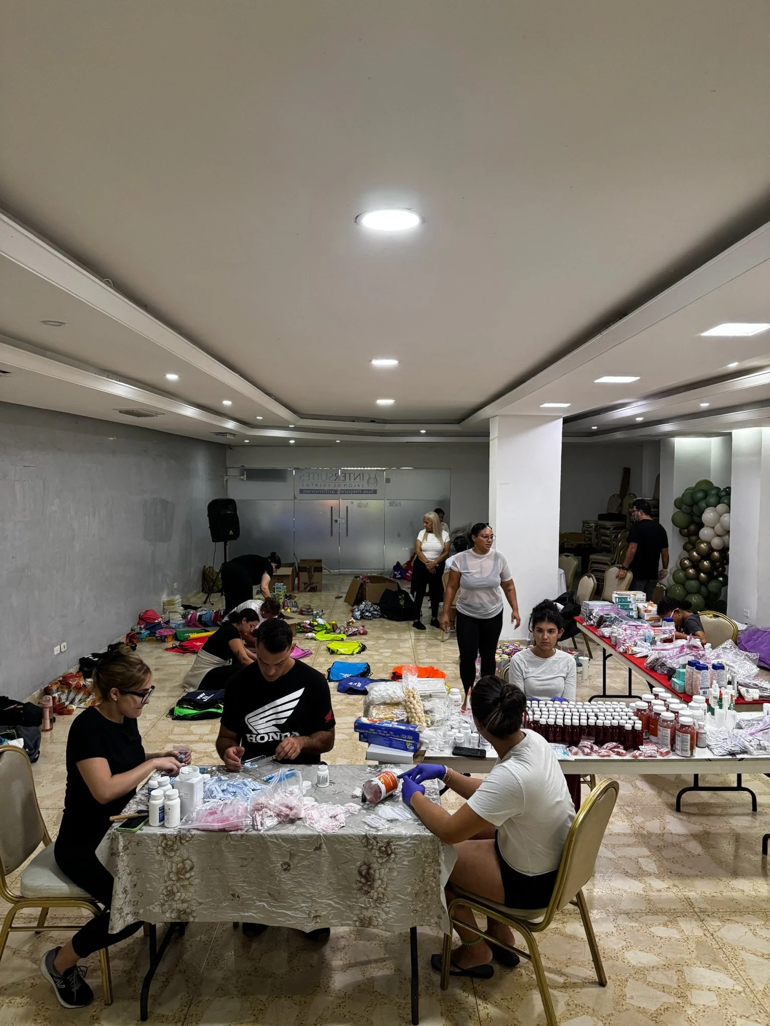 People working on organized table of supplies and items in a large, well-lit indoor space with balloons and chairs in the background.
