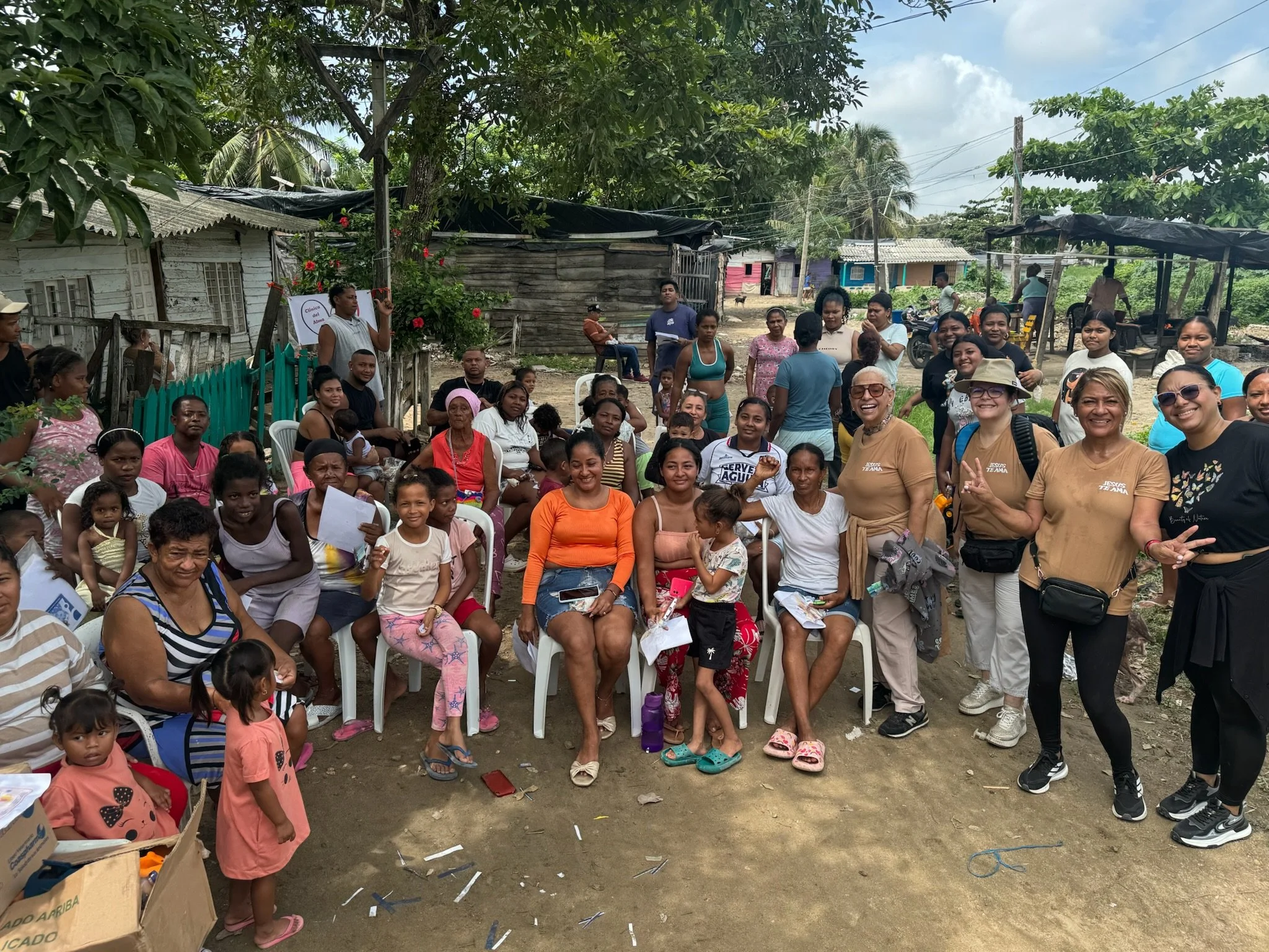 Group of people, including children and adults, gathered outdoors in a rural area, some sitting on white plastic chairs, others standing, with trees and simple houses in the background.
