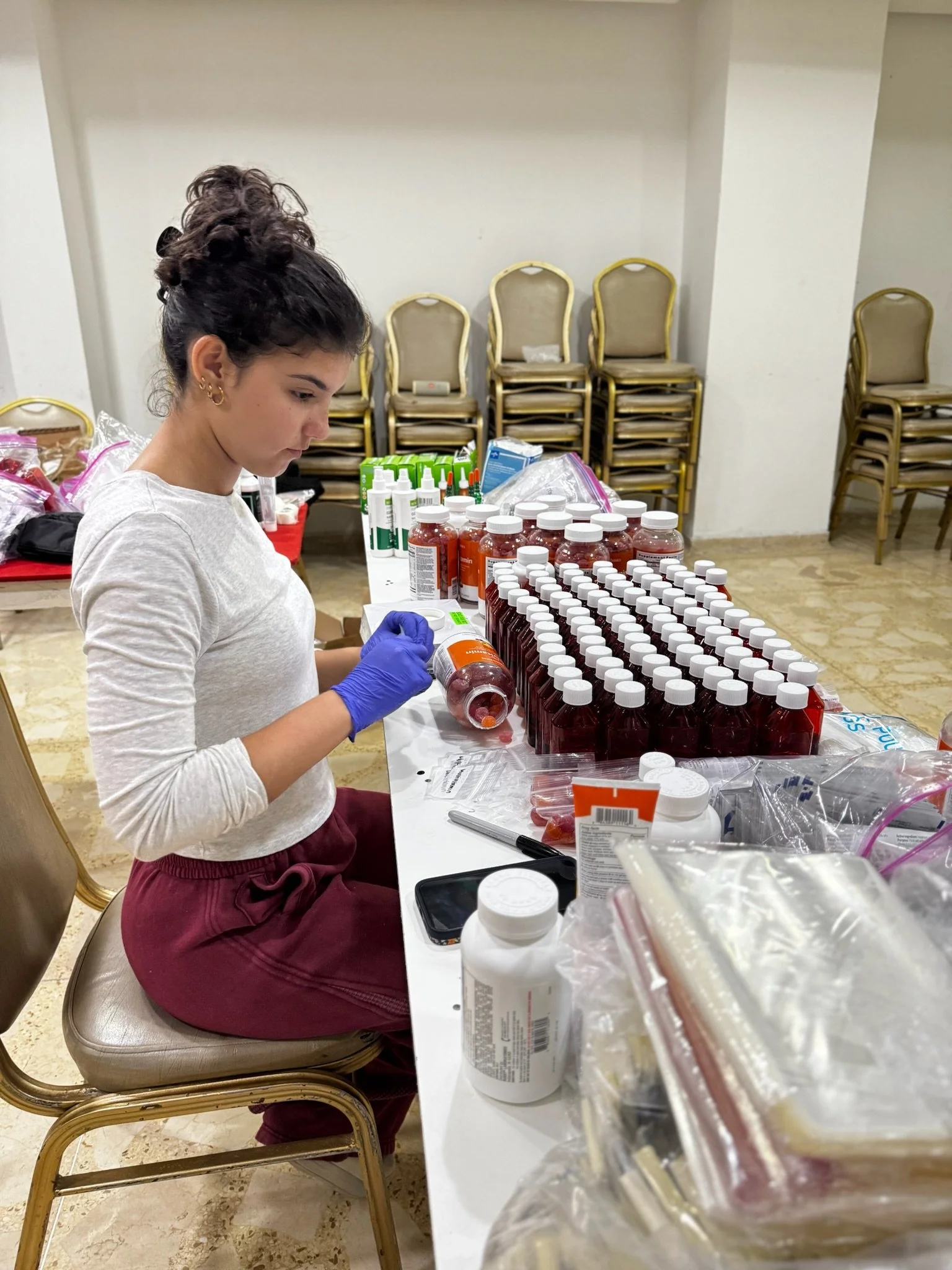 A woman wearing purple gloves is organizing medication bottles at a table covered with medical supplies, with stacked chairs in the background.