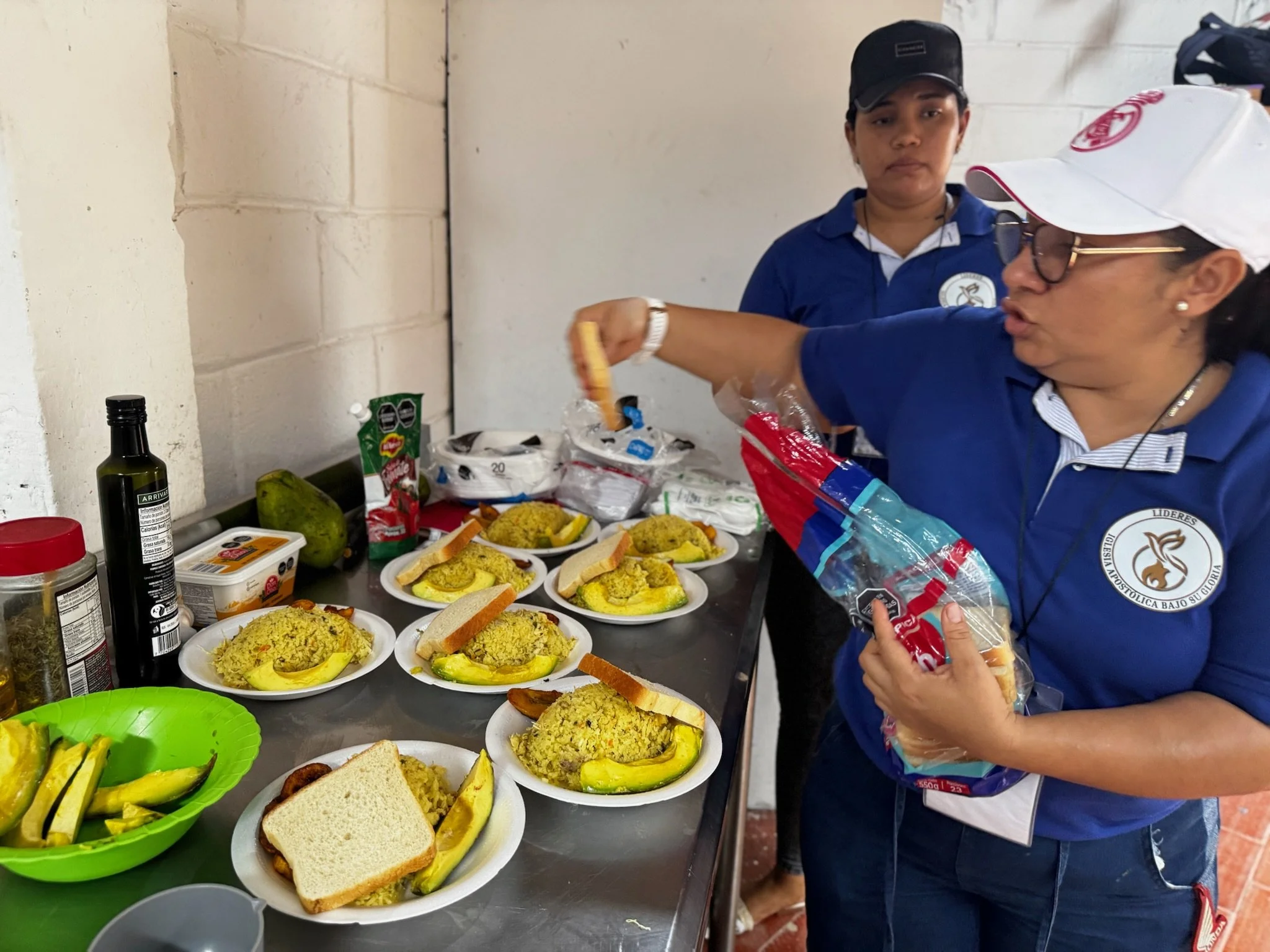 Two women in blue shirts and hats preparing food on a kitchen table. The table has plates with food, including bread and plantains, and various ingredients and supplies.