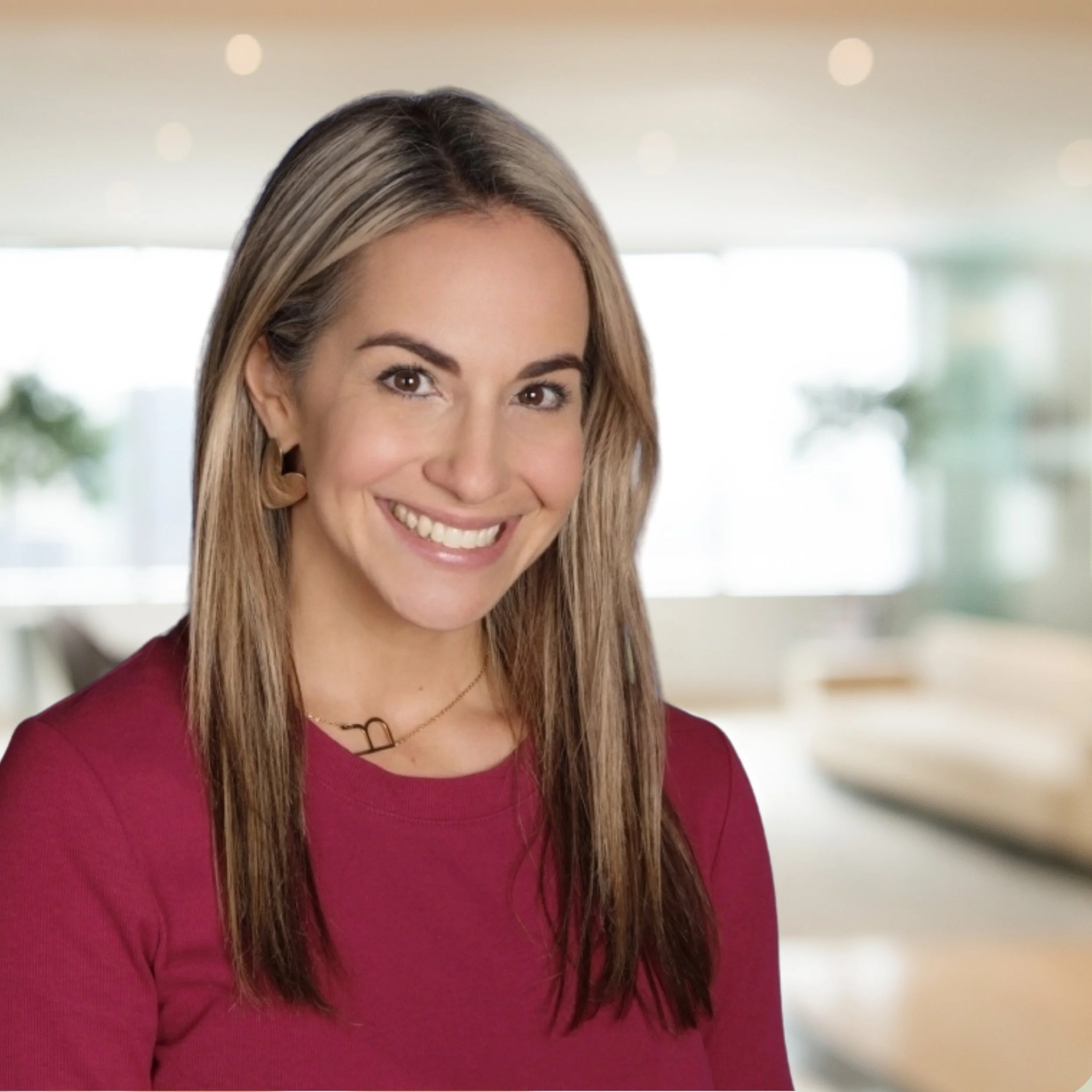 A smiling woman with long brown hair wearing a burgundy top and earrings, standing in a bright, modern living room with large windows and blurred furniture in the background.