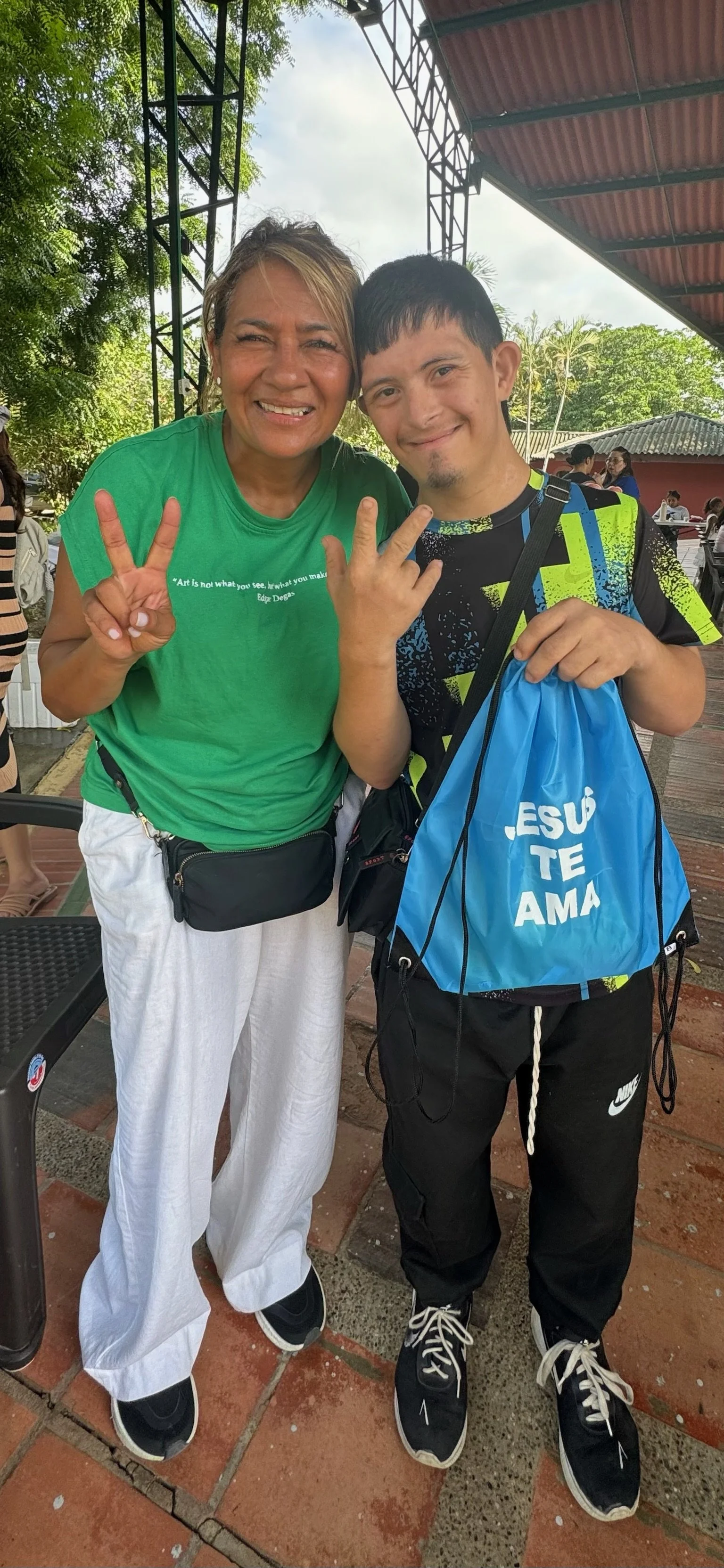 A woman and a young man smiling and making peace signs at an outdoor event, with trees and a roofed structure in the background.
