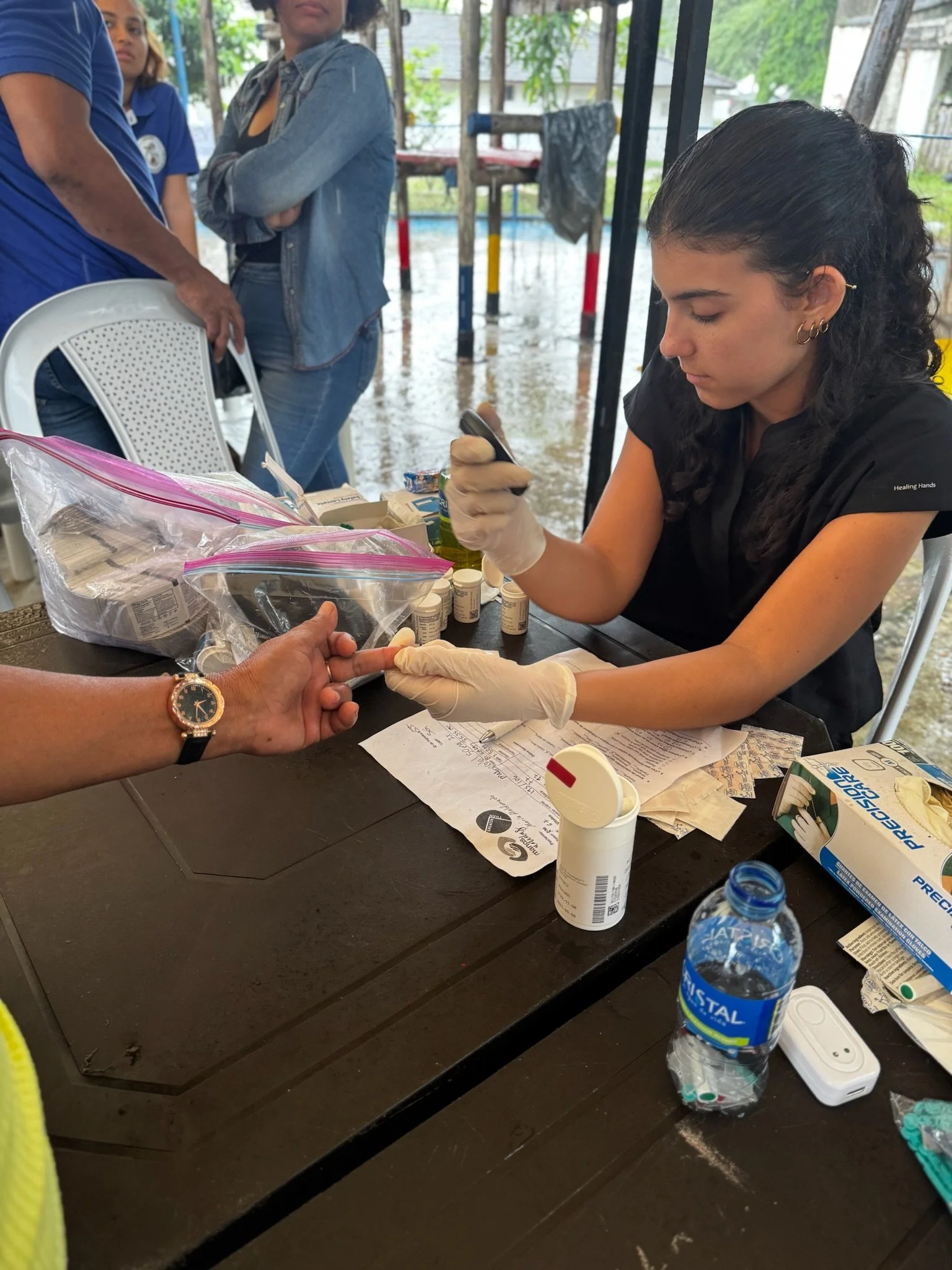 A woman wearing gloves is drawing blood from a person's finger during a medical testing procedure at a table filled with medical supplies. In the background, several people are standing outside in a rainy setting.