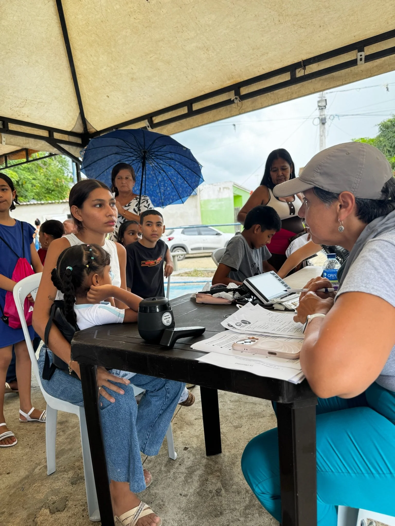 A group of children and adults at an outdoor health or registration event, with a woman sitting at a table with documents, speaking to a young girl sitting on a woman's lap.