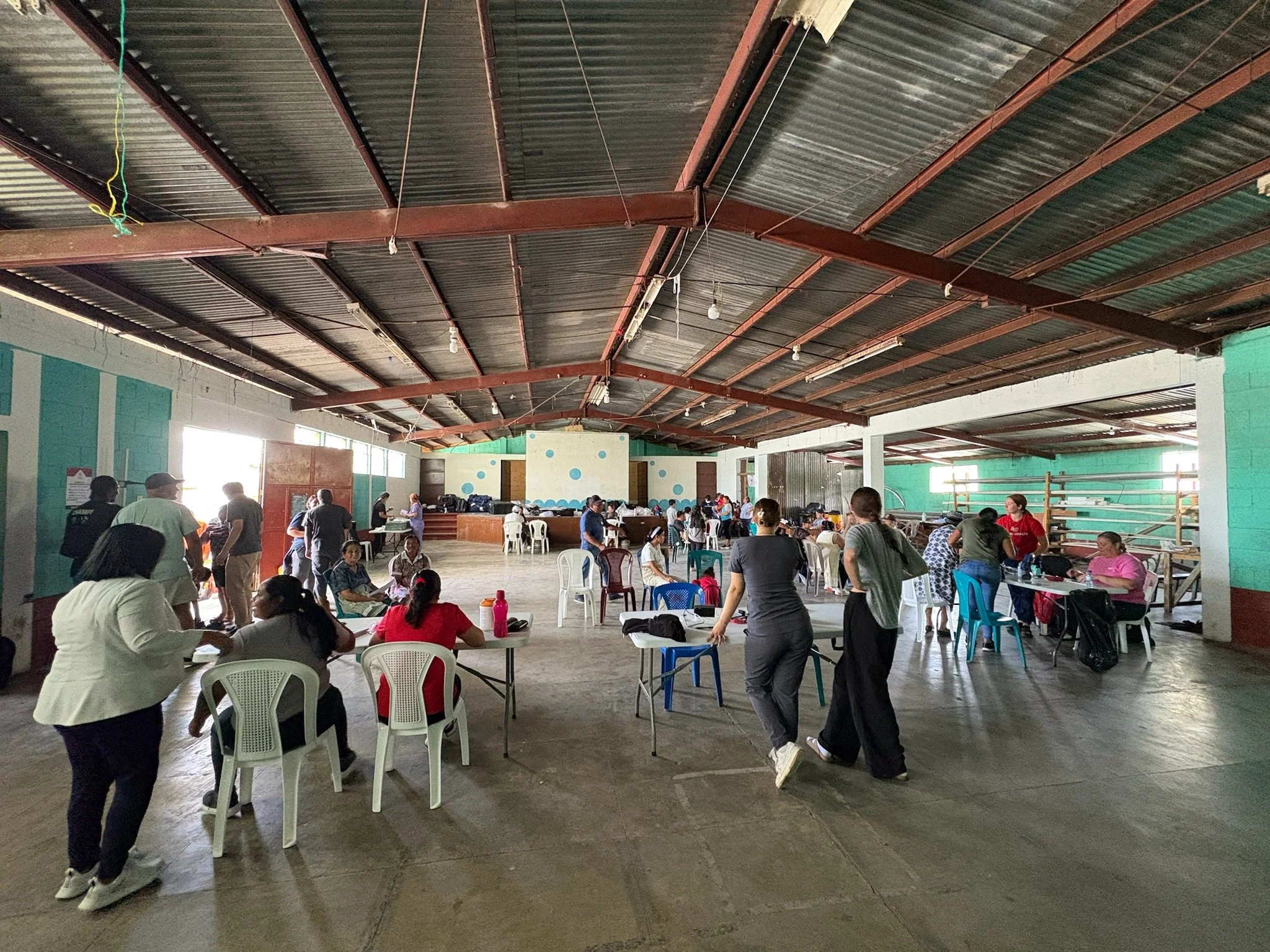 People gathering inside a large open hall with a high metal roof, some seated at tables, others standing and walking around.