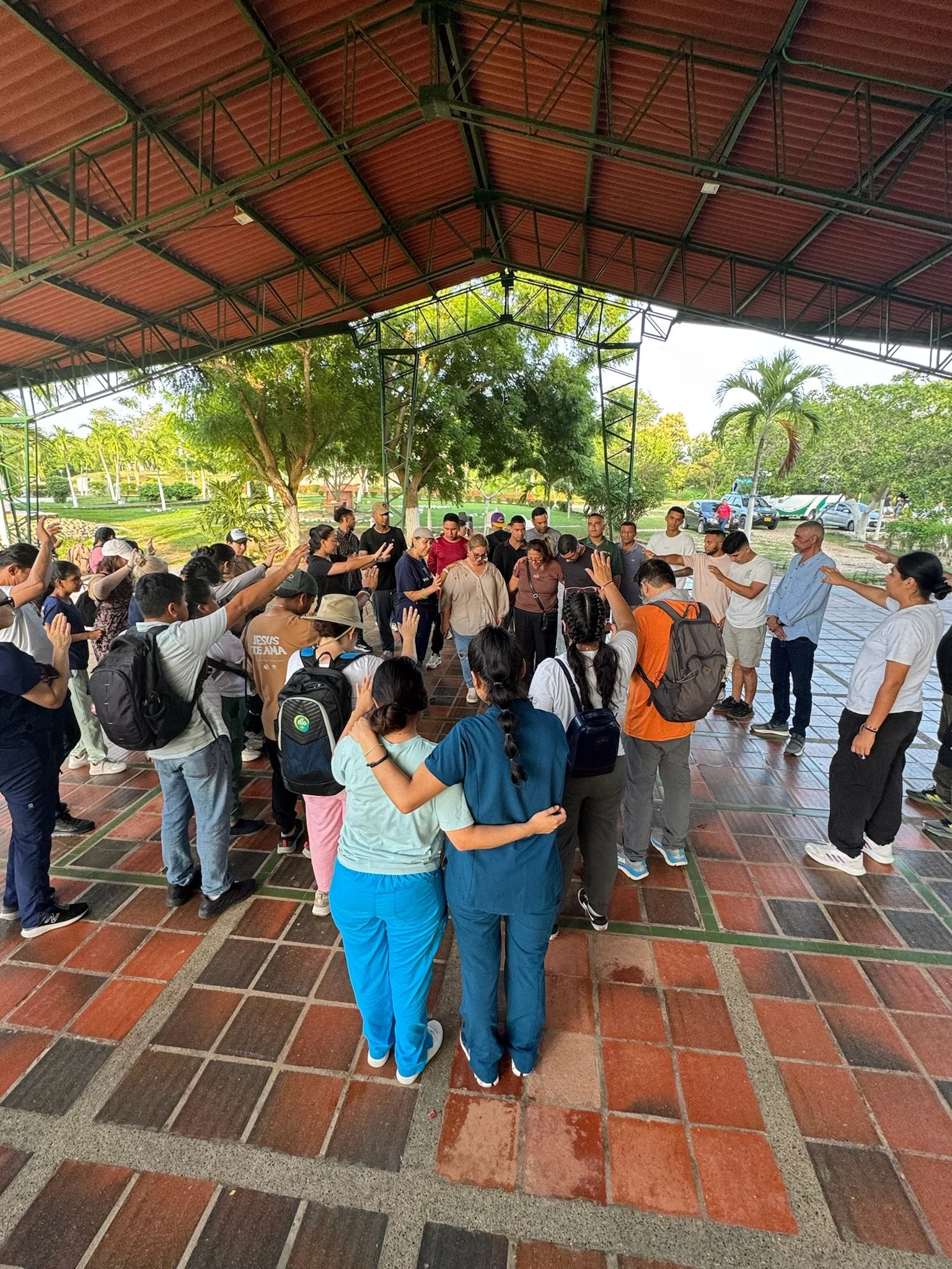 A group of people gathered under a large, open-sided pavilion with red roof tiles, some with hands raised in prayer or reflection. The setting is outdoors with trees and parked cars visible in the background.