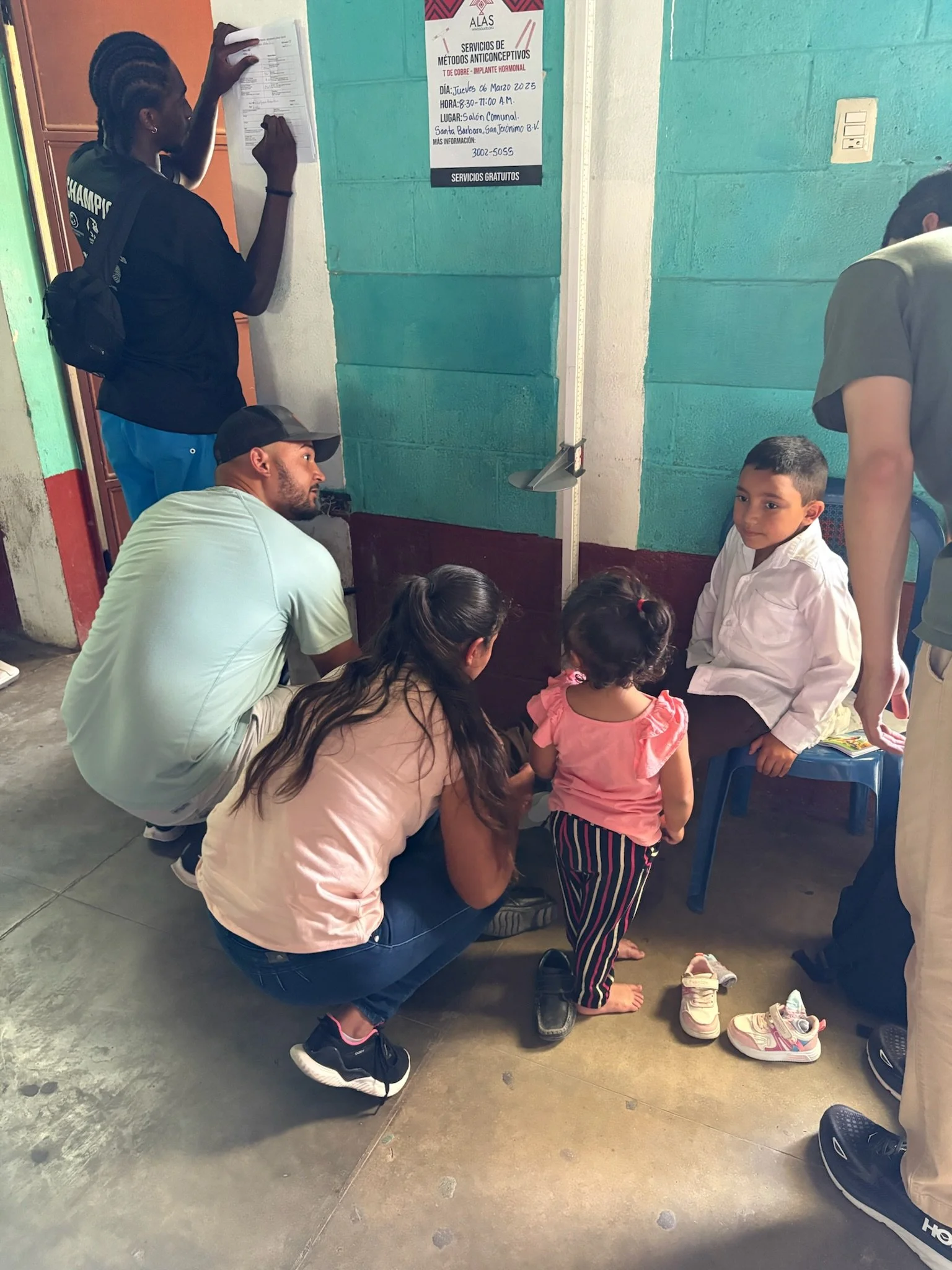 A young girl is having her health checked by volunteers or healthcare workers in a community health setting. Others are sitting or standing nearby, and shoes are on the floor.