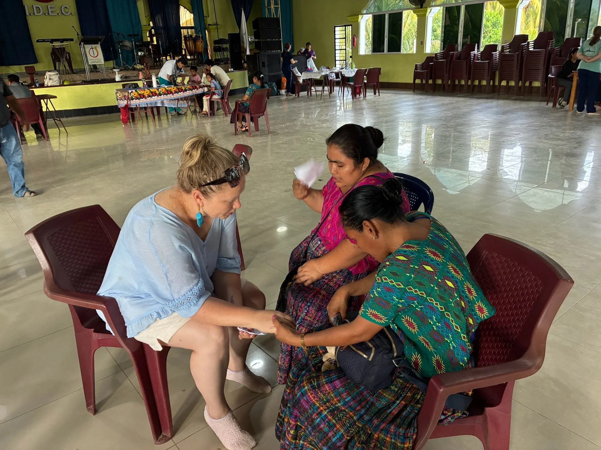Three women sitting in a circle at a community event in a large hall, with colorful surroundings and other people in the background.