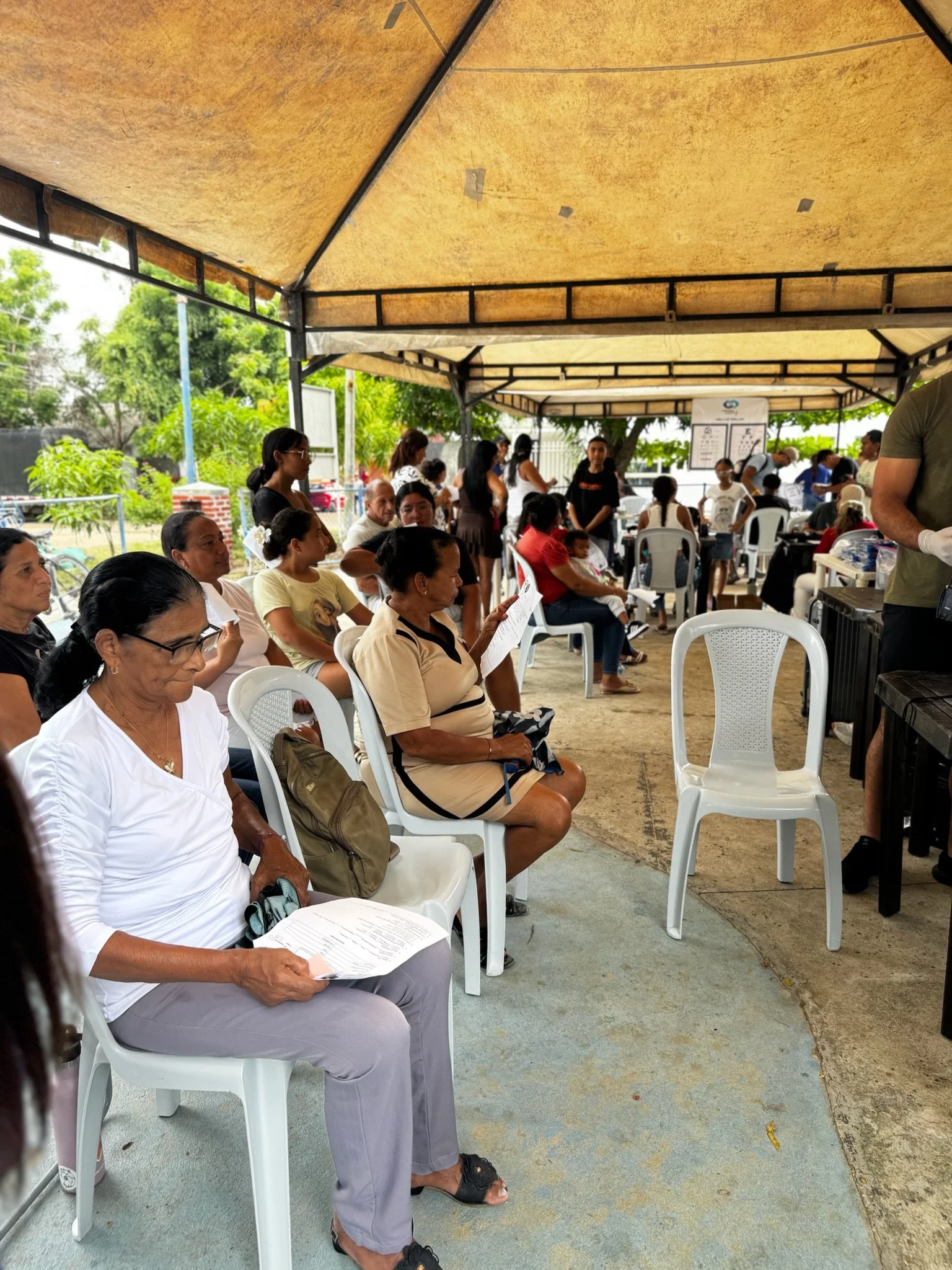 People seated in a shaded outdoor area, waiting in line or for an event, with some reading papers and others talking, under a canopy with trees and greenery in the background.