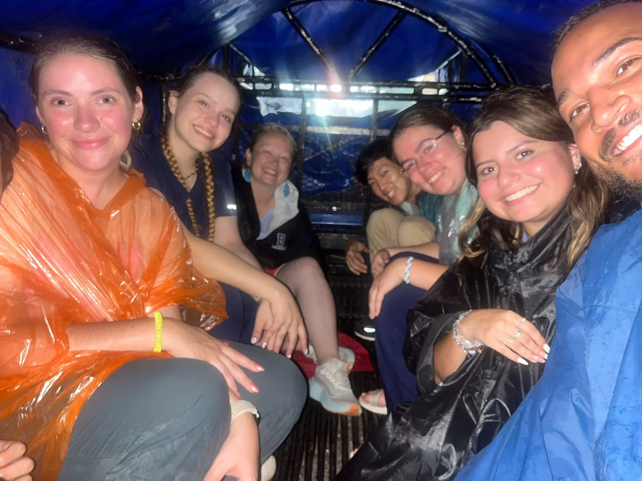 Group of smiling people sitting closely inside a blue tent or structure, some wearing rain ponchos, with daylight visible in the background.