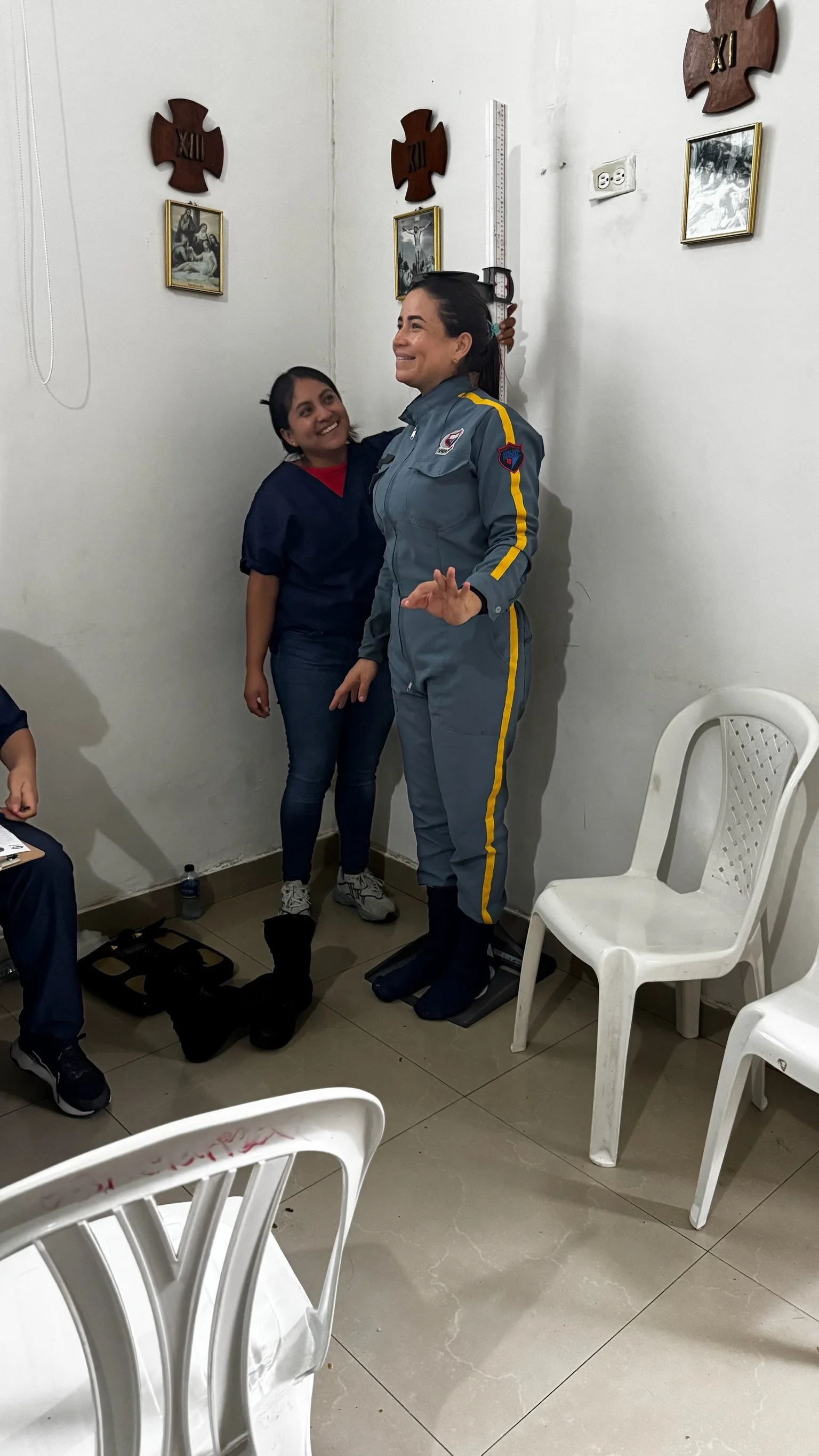 A woman in a gray police uniform standing on a weighing scale with her arm around a young girl, both smiling in a room with white walls and chairs.