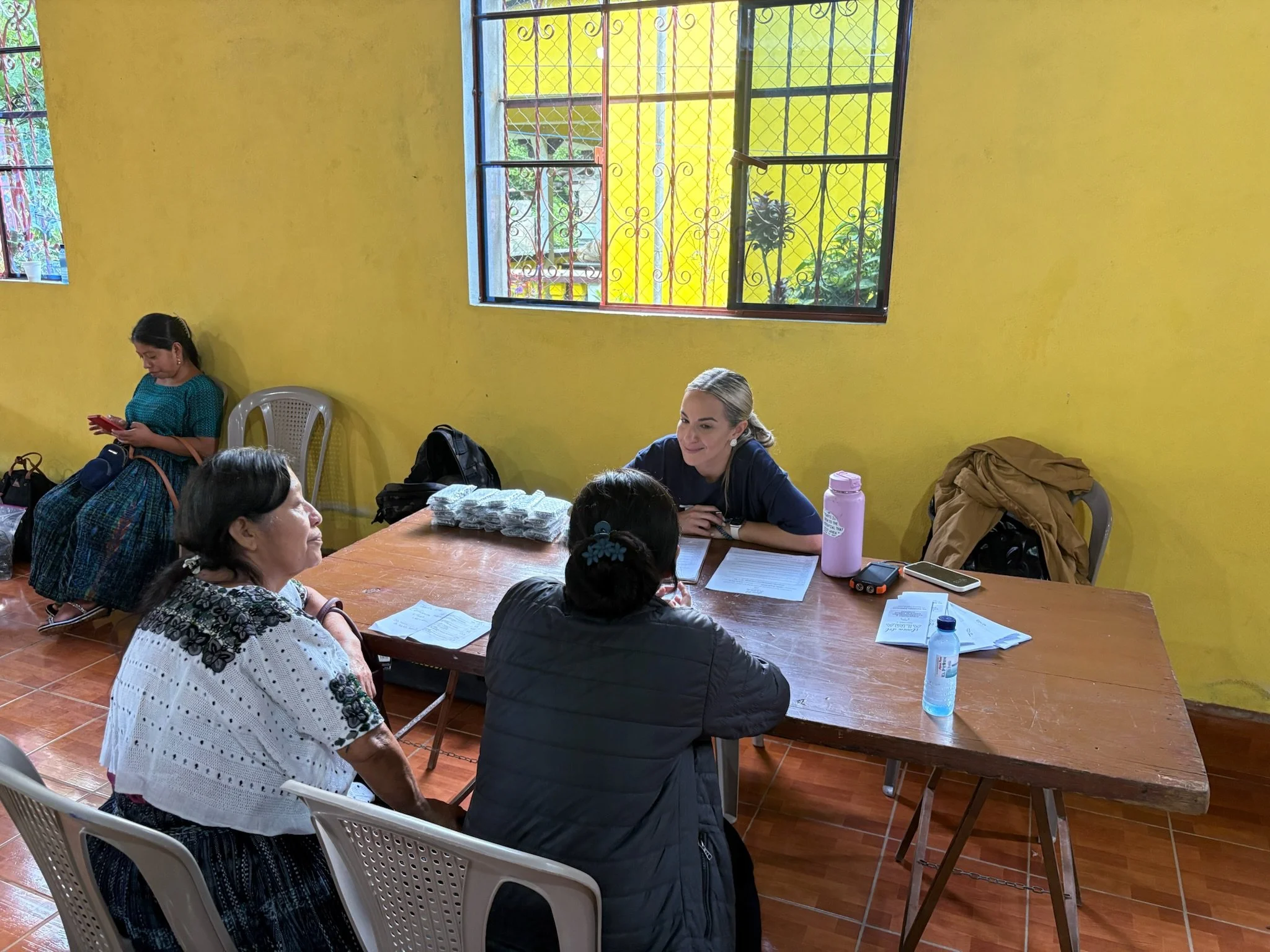 Four women sitting around a wooden table in a room with yellow walls and barred windows. One woman is sitting at the far end of the table, smiling, with papers and a pink water bottle in front of her. Two women are sitting next to her, having a conve