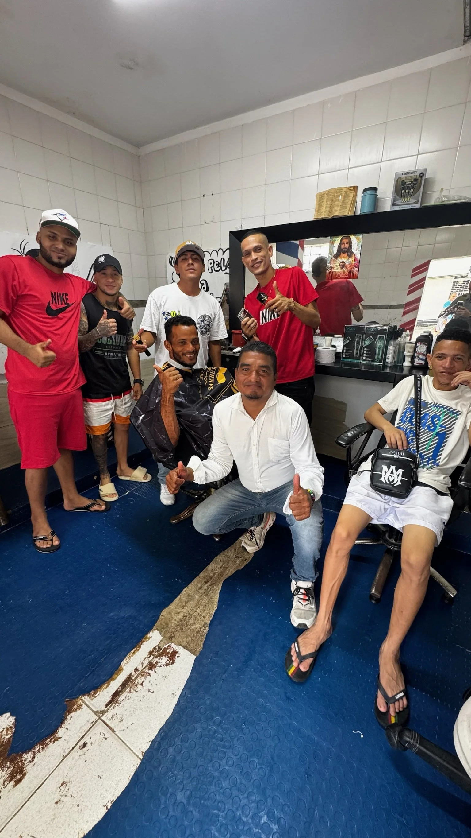 Group of men inside a barbershop, some sitting, some standing, with hair and barber tools behind the mirror.