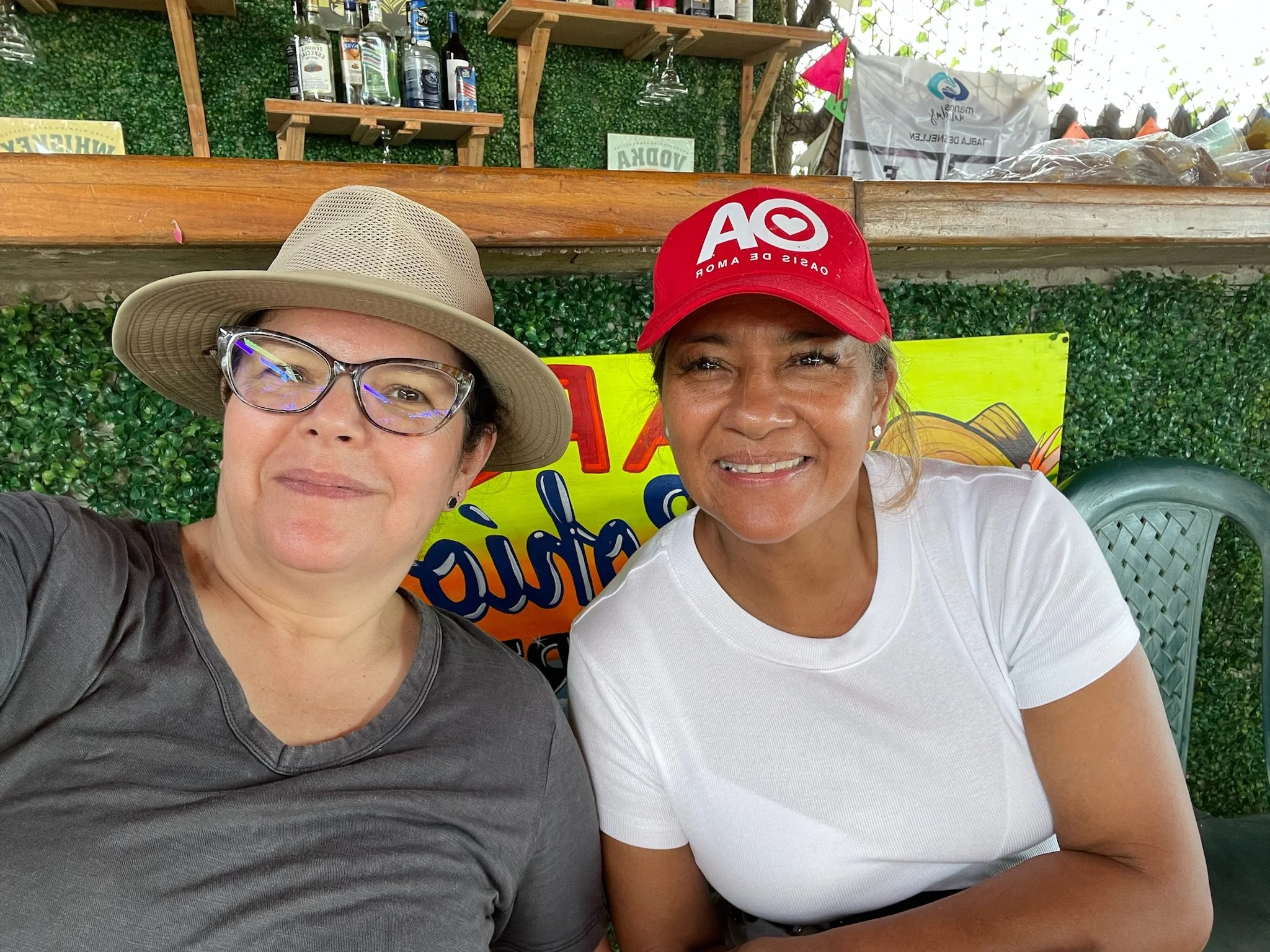 Two women sitting outdoors at a table, smiling for a selfie. The woman on the left has short dark hair, glasses, a tan wide-brimmed hat, and a dark gray shirt. The woman on the right has dark hair pulled back, a red cap with a white logo, earrings, a