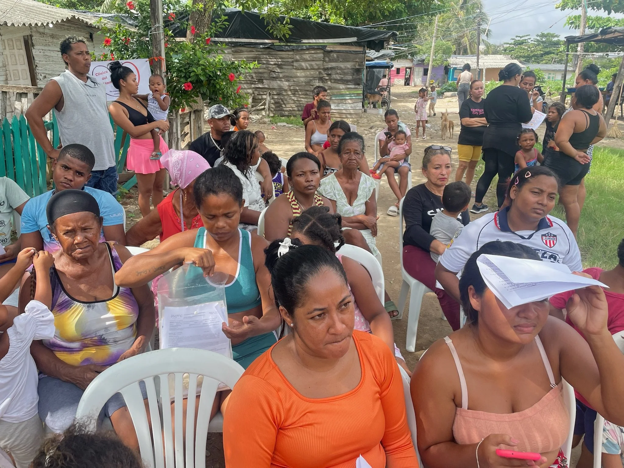 A group of people gathered outdoors, some seated and some standing, in a rural area with simple houses and trees in the background. They appear to be attending a community event or meeting.