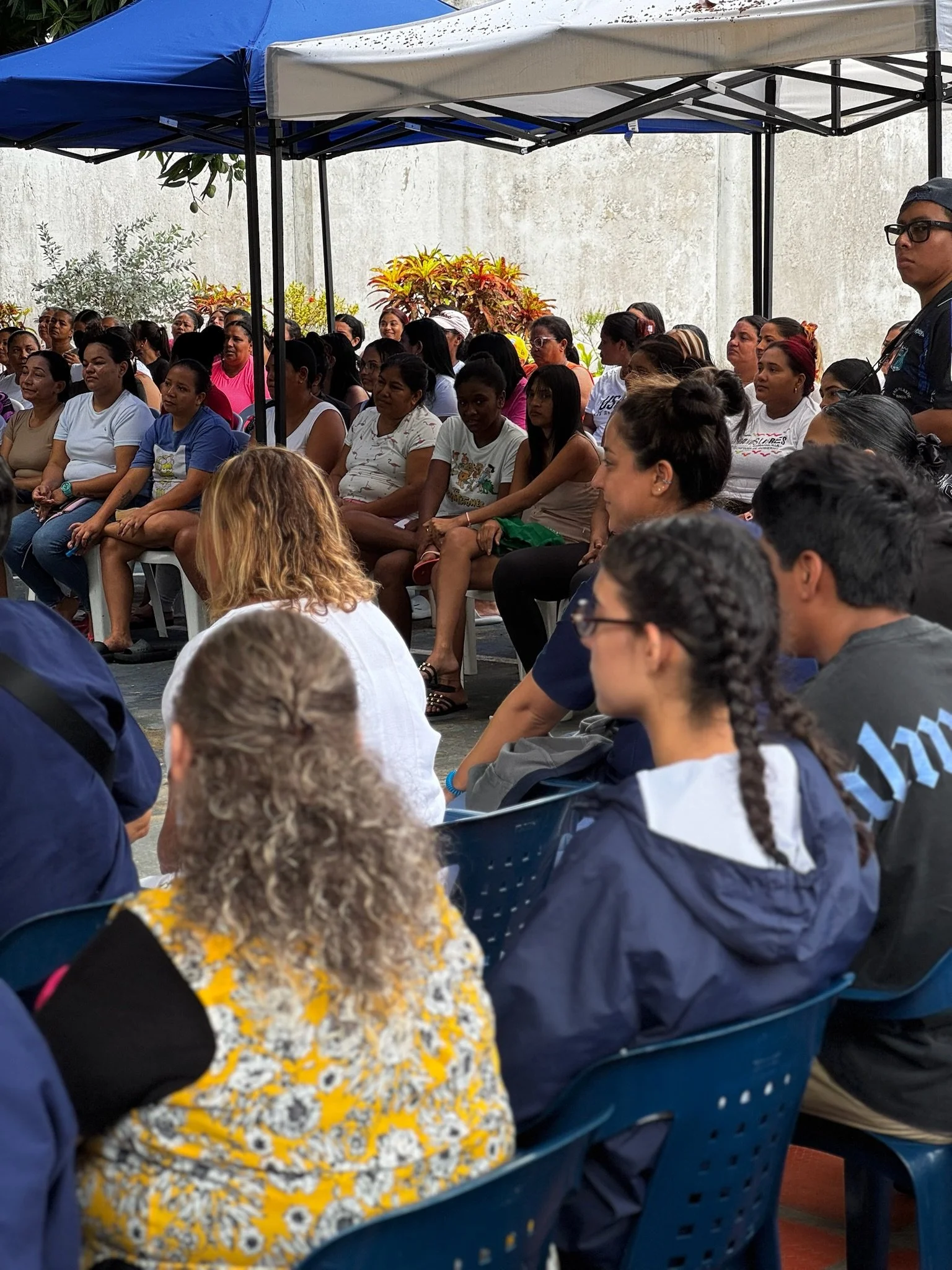 Crowd of people seated under canopy at outdoor event, some are watching attentively.