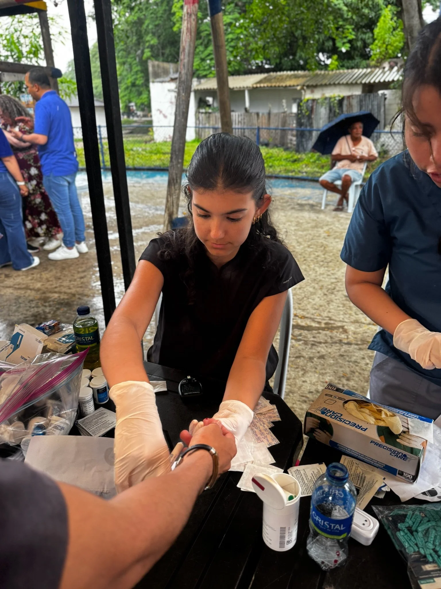 A young woman receiving medical assistance during a health event, with healthcare supplies on the table, outdoors on a rainy day.