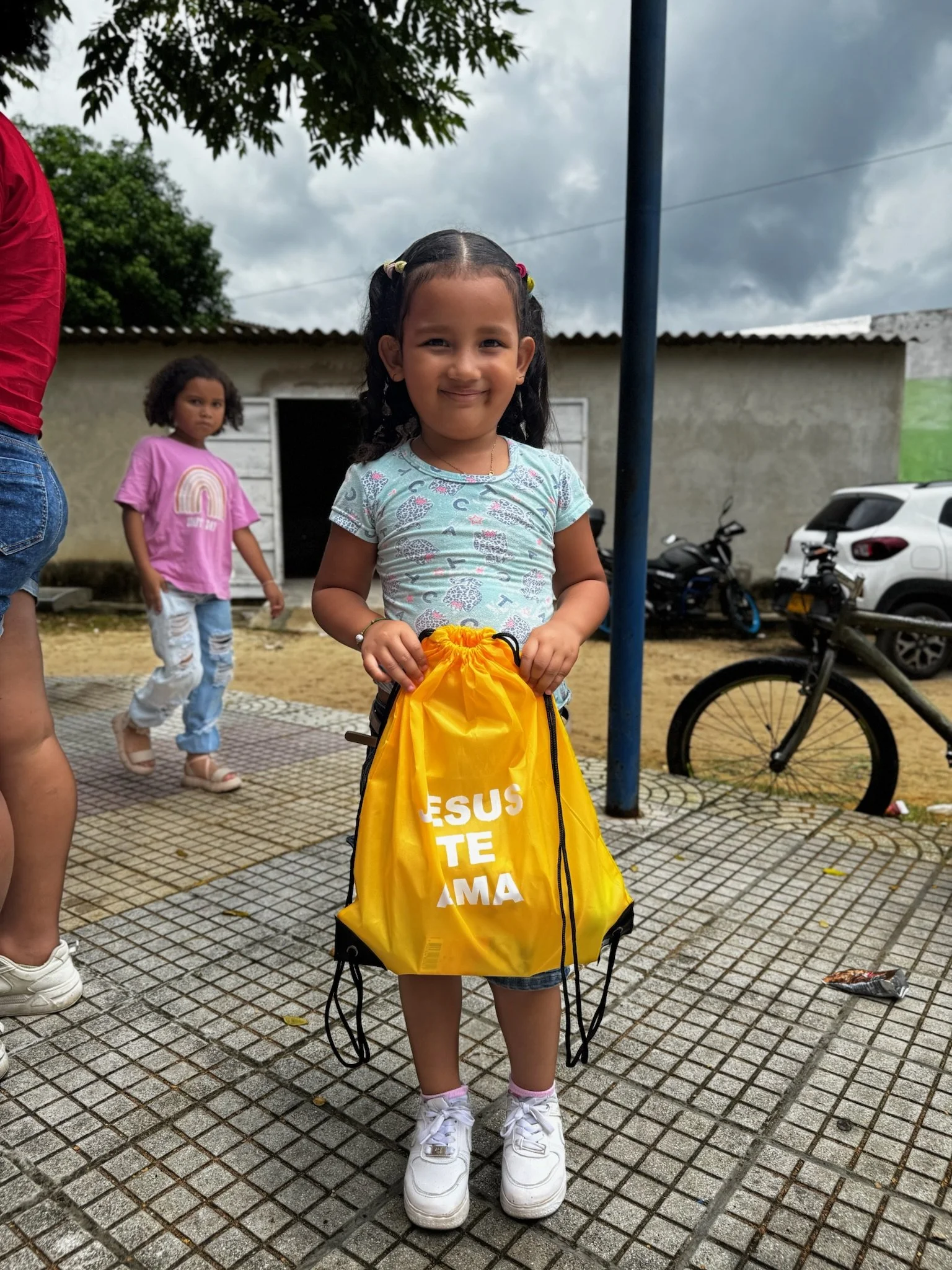 A young girl with dark hair in pigtails, wearing a light blue shirt with patterns and denim shorts, holding a yellow bag with the words 'JESUS TE AMA' in white, standing outdoors on a tiled surface. Behind her are two other children, a woman with sho