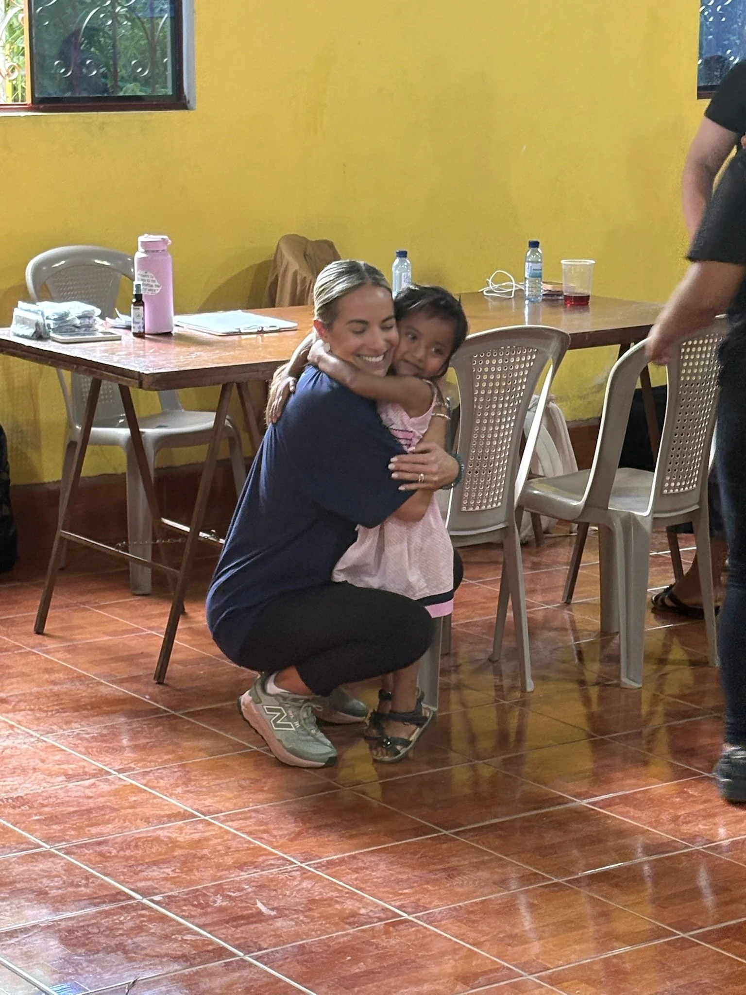 A woman kneeling down and hugging a young girl in a room with yellow walls and tiled floor.