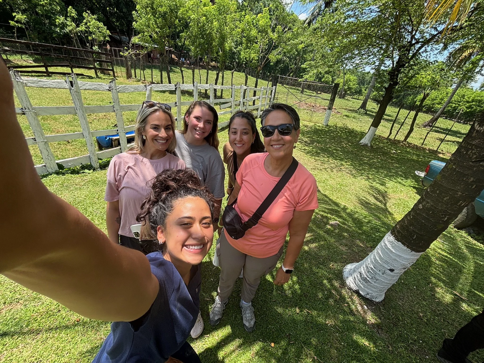 Group of six smiling women taking a selfie outdoors on a sunny day, standing on green grass near trees and a white wooden fence.