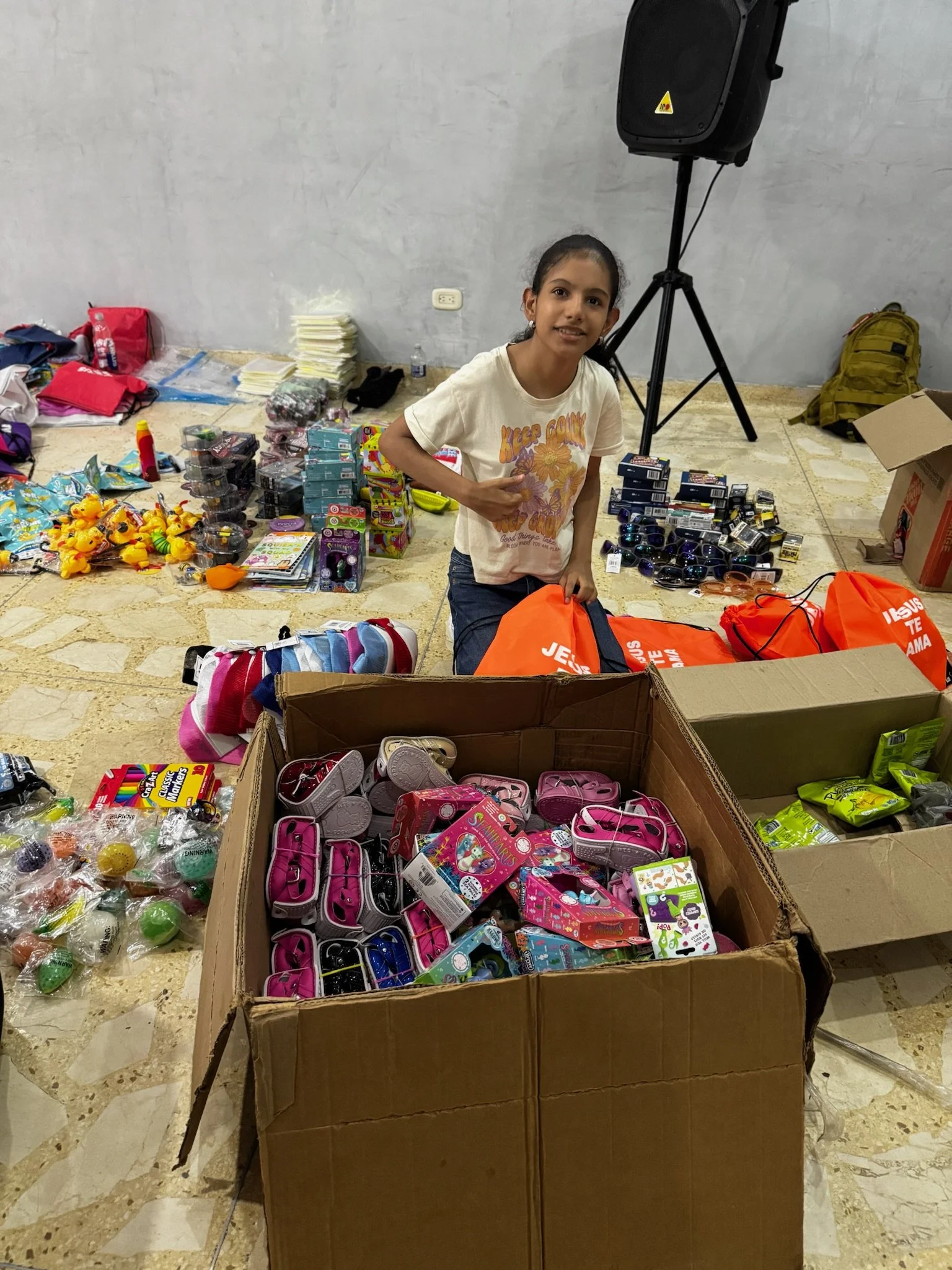 Young girl surrounded by toys and school supplies, sitting behind a large cardboard box filled with pink and purple toy backpacks. Various toys, sunglasses, and other items are arranged on the ground around her. In the background, there are additiona