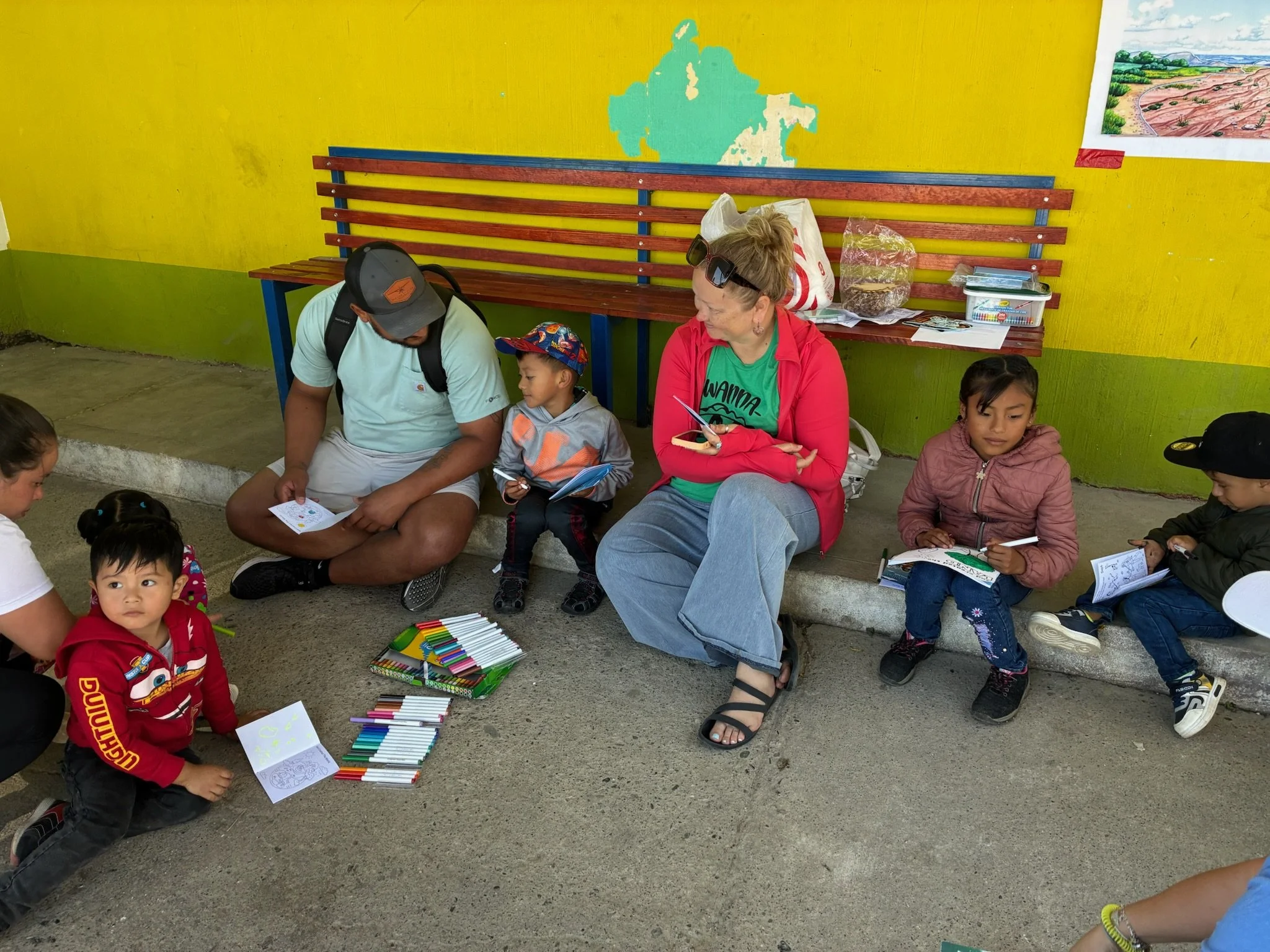 Children and two adults sitting on the ground and on a low concrete ledge, engaging in coloring activities. The kids have coloring books and markers in front of them, and a woman in a red jacket and sandals is reading or writing in a notebook. The ch