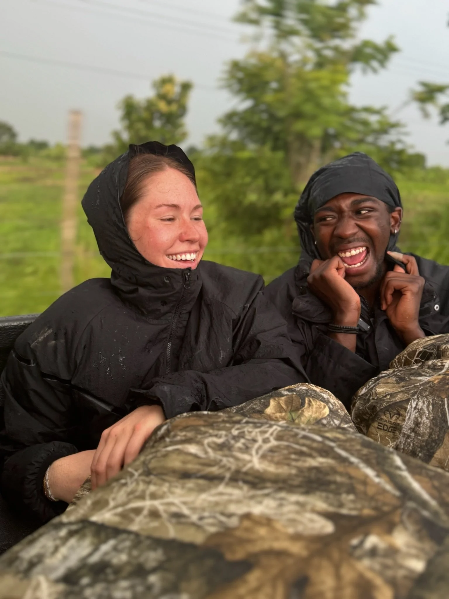 Two people in rain jackets laughing and enjoying an outdoor activity, with trees and greenery in the background.