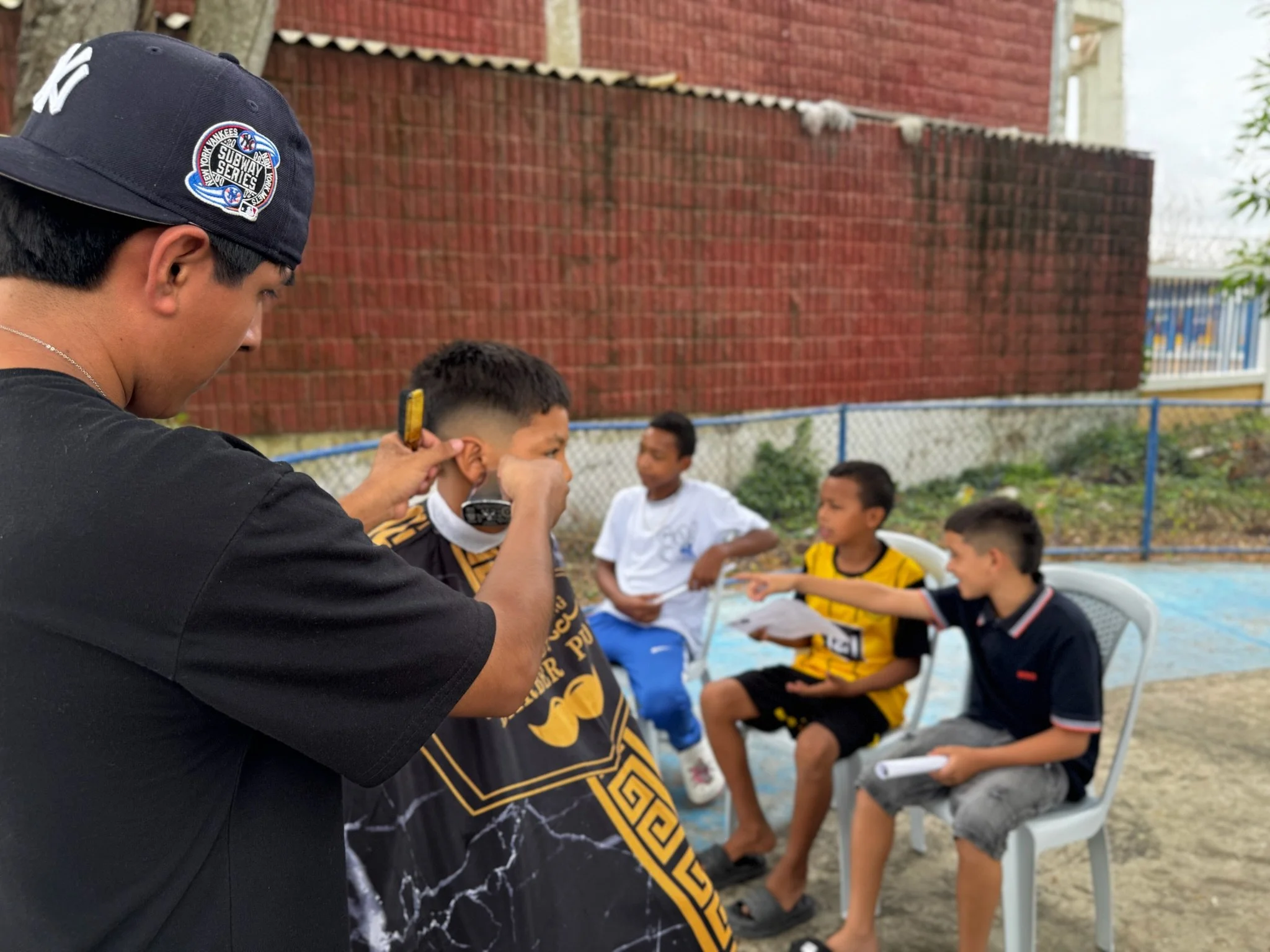 A young man is grooming a boy wearing a black and gold scarf, while three other boys sit on plastic chairs nearby, with one of them pointing and engaged in conversation during an outdoor gathering.