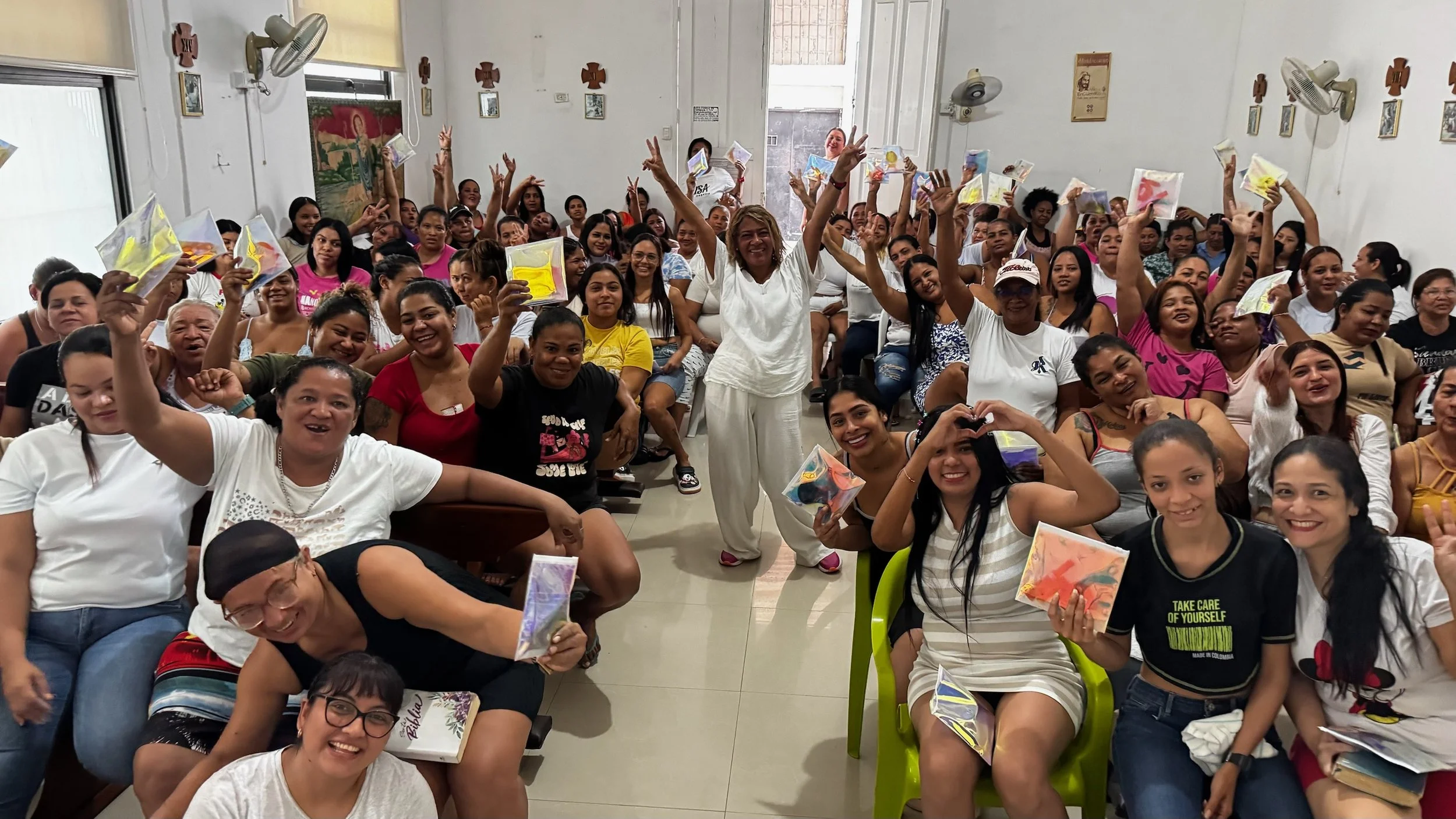 A large group of smiling women in a classroom, some holding colorful envelopes or packages, with a woman standing in the center raising her hands and surrounded by others celebrating.