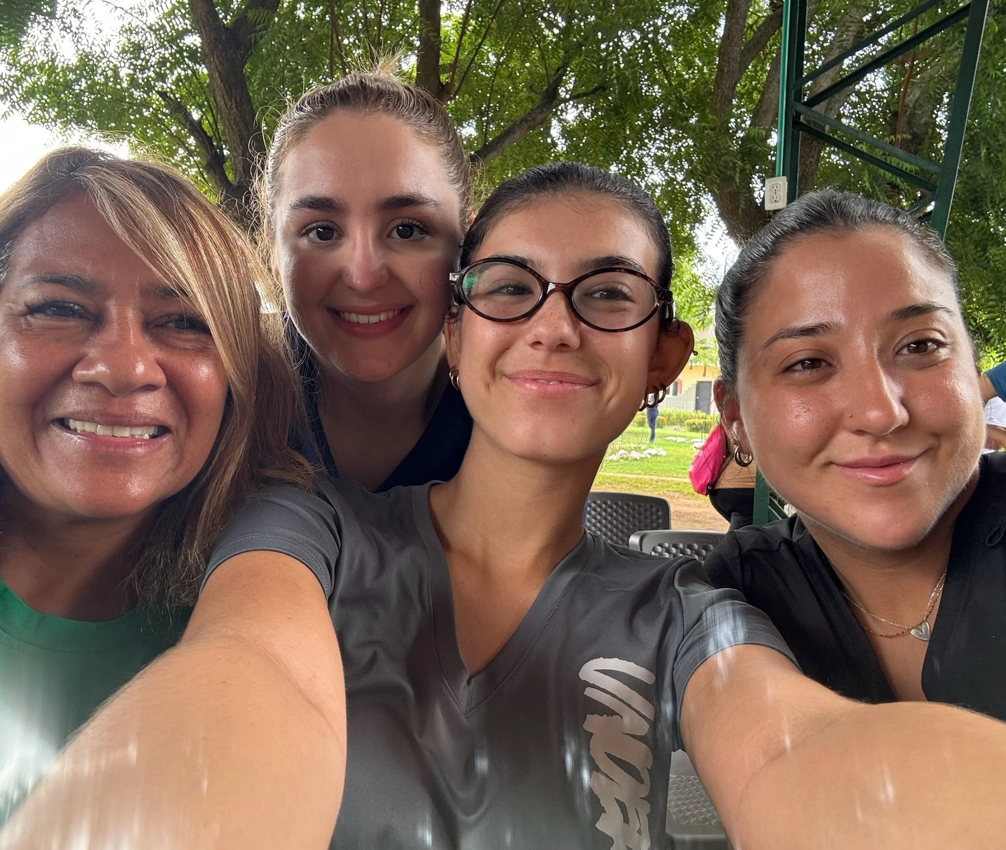 Four women smiling, taking a selfie outdoors under a tree.