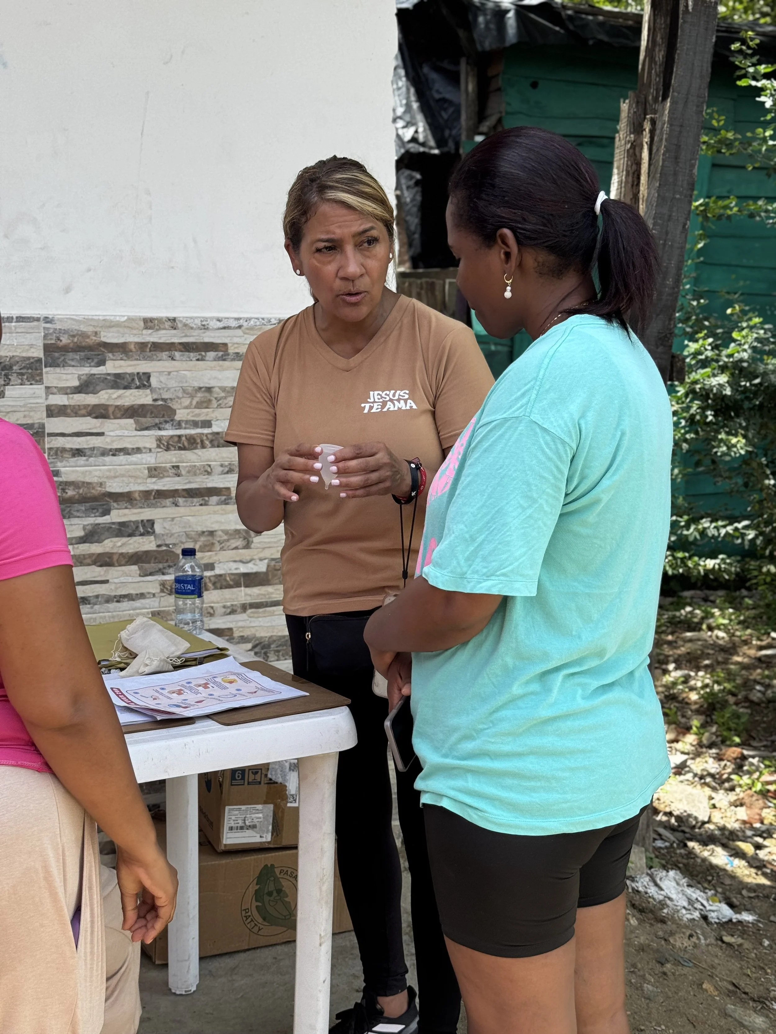 Two women are engaged in a conversation outside, near a table with some papers, a water bottle, and a cardboard box underneath. One woman wears a tan t-shirt with white text, and the other wears a light blue t-shirt. They are standing close to each o