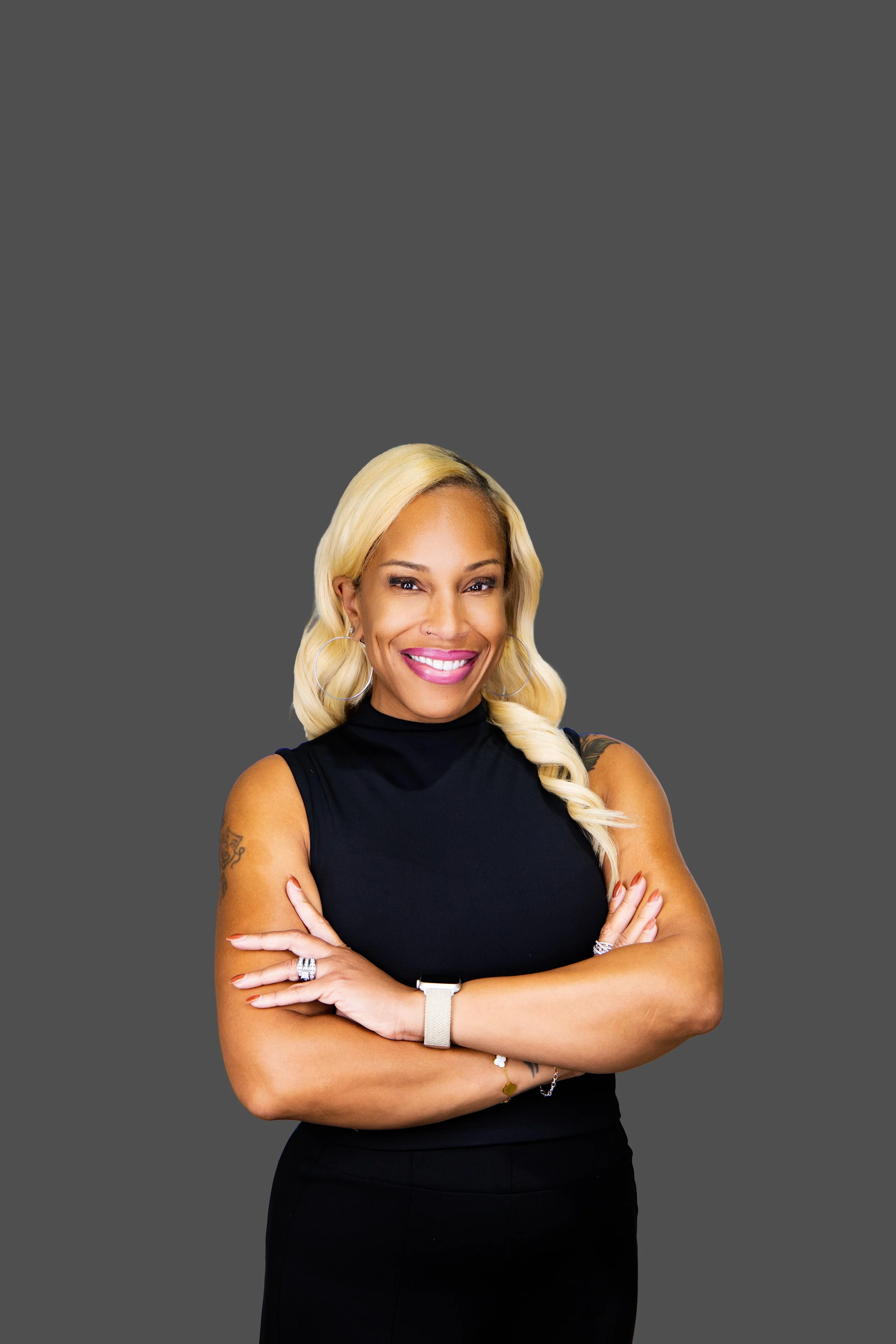A woman with blonde hair styled in loose waves, wearing hoop earrings, a black sleeveless top, and a watch, standing with crossed arms and smiling against a gray background.
