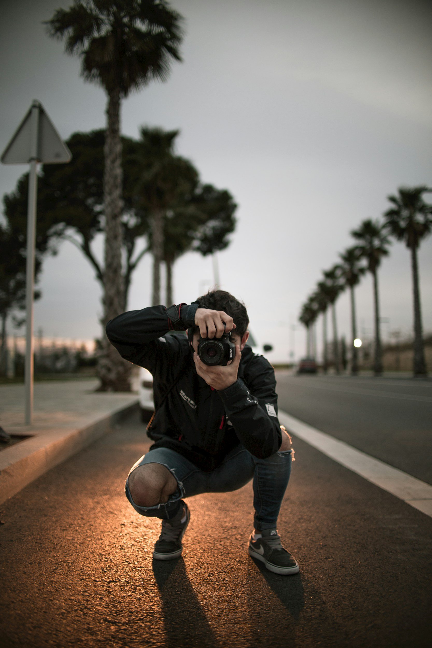 A person crouching on a street at dusk, taking a photo with a camera. The background features tall palm trees and a cloudy sky.