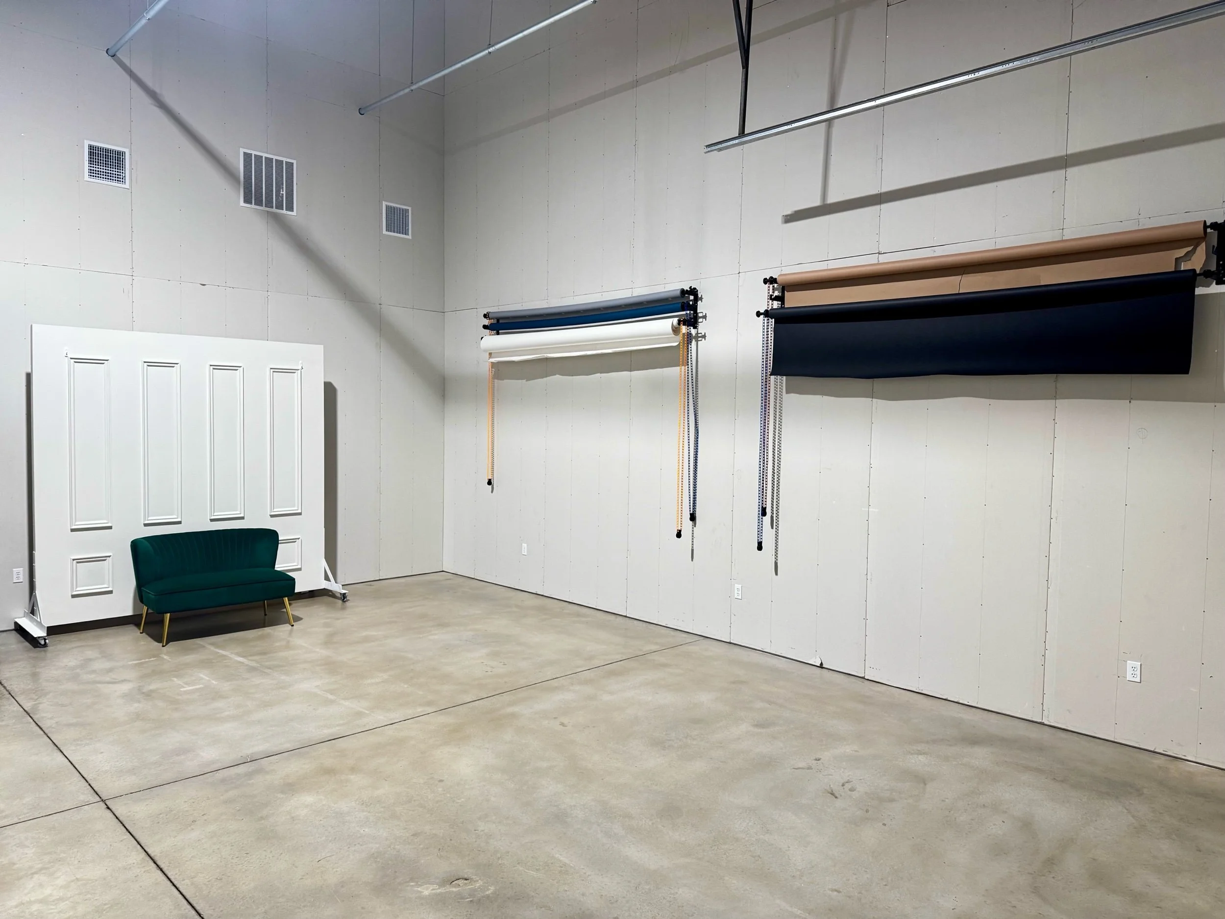 Empty photography studio with white wall backdrop, green velvet sofa, and three rolls of backdrop paper in different colors hanging on the wall.