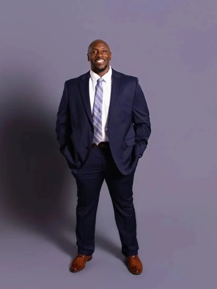 A man in a navy suit, white shirt, and patterned tie standing against a gray background, smiling with hands in his pockets.