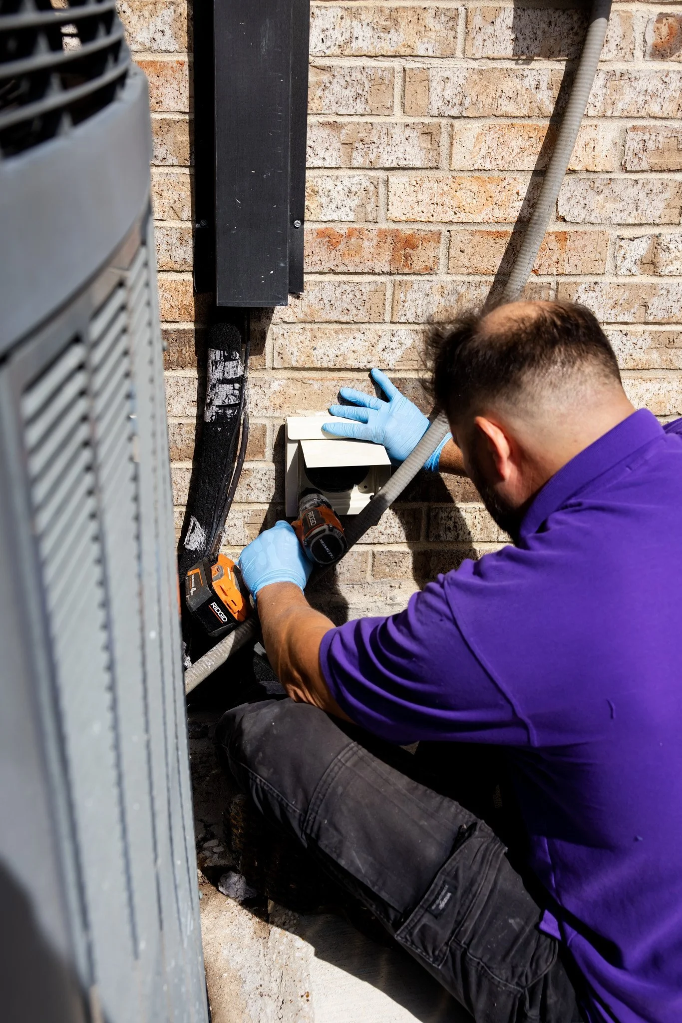 A technician working on an outdoor electrical box, wearing blue gloves and using a cordless drill, with a brick wall and a utility pipe in the background.