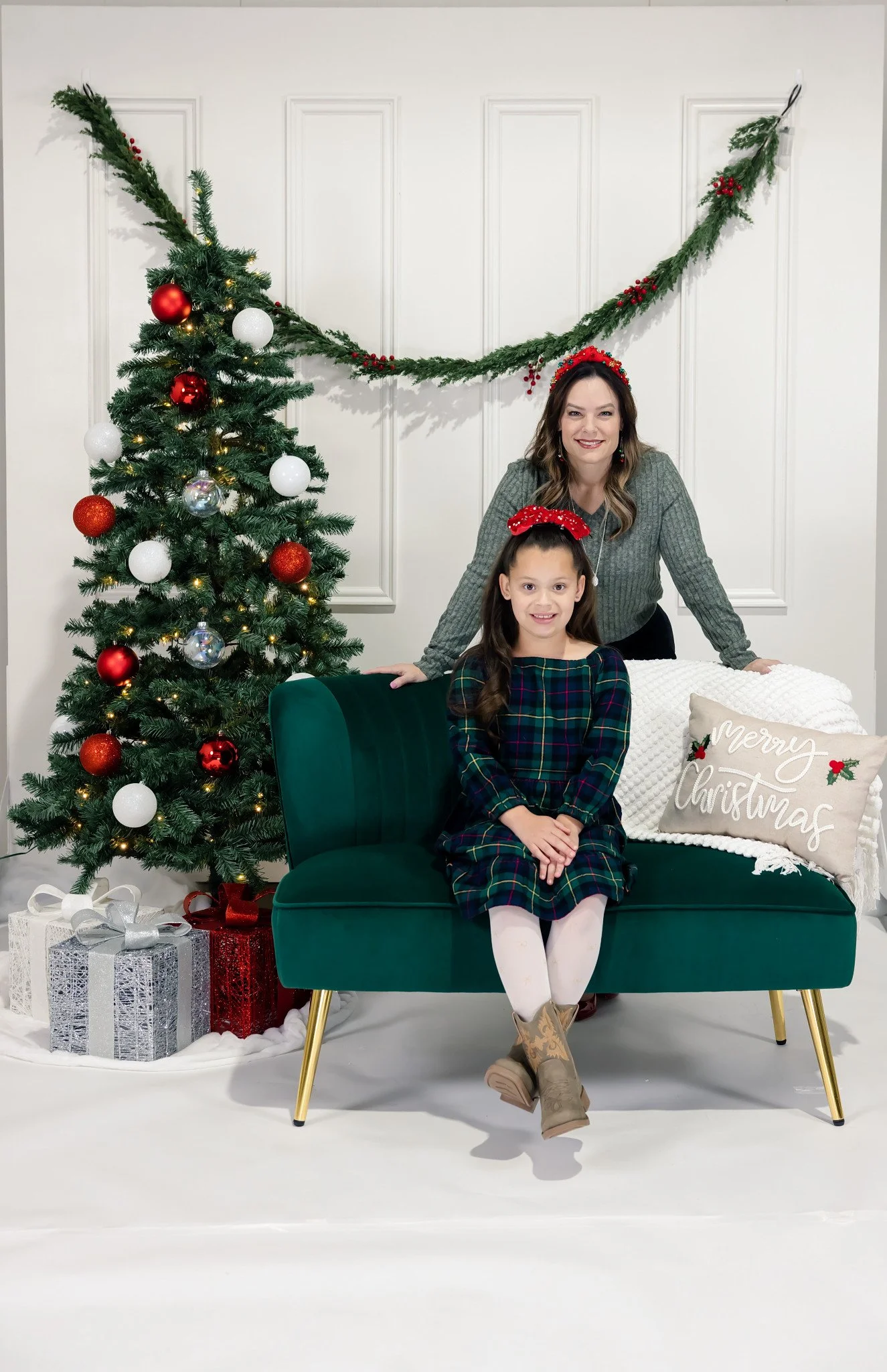 A woman and a girl sitting and standing on a teal sofa with gold legs in a holiday scene. A decorated Christmas tree with red, white, and silver ornaments is to their left. Presents wrapped in silver, white, and red with ribbons are under the tree. A white throw pillow on the sofa reads 'Merry Christmas'. The woman and girl are wearing red holiday headbands with berries. The background has a white wall with holiday greenery and red berries hanging.